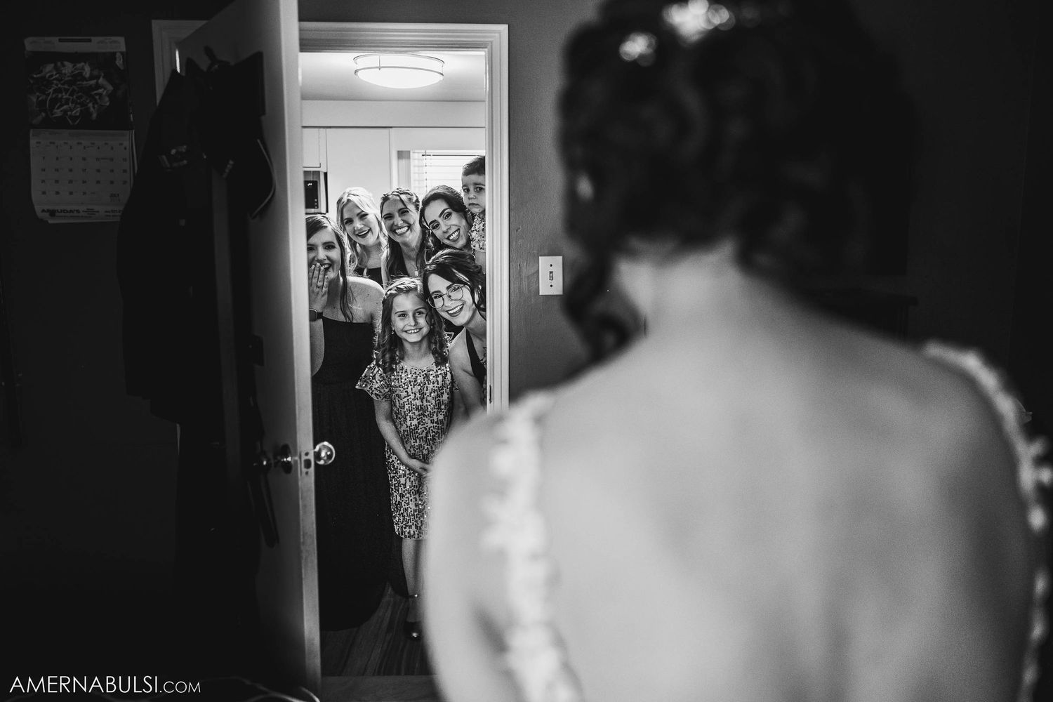 Black and white image of bridesmaids peeking through doorway at wedding preparations.