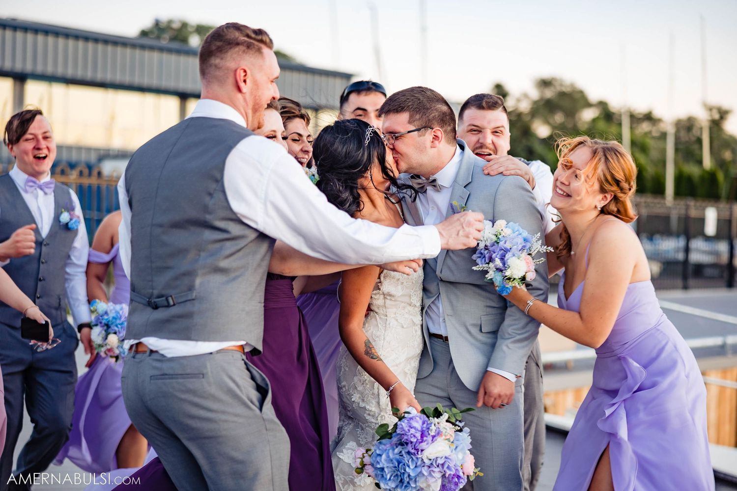 Playful wedding party shares an energetic moment on a deck with bridesmaids in lavender dresses.