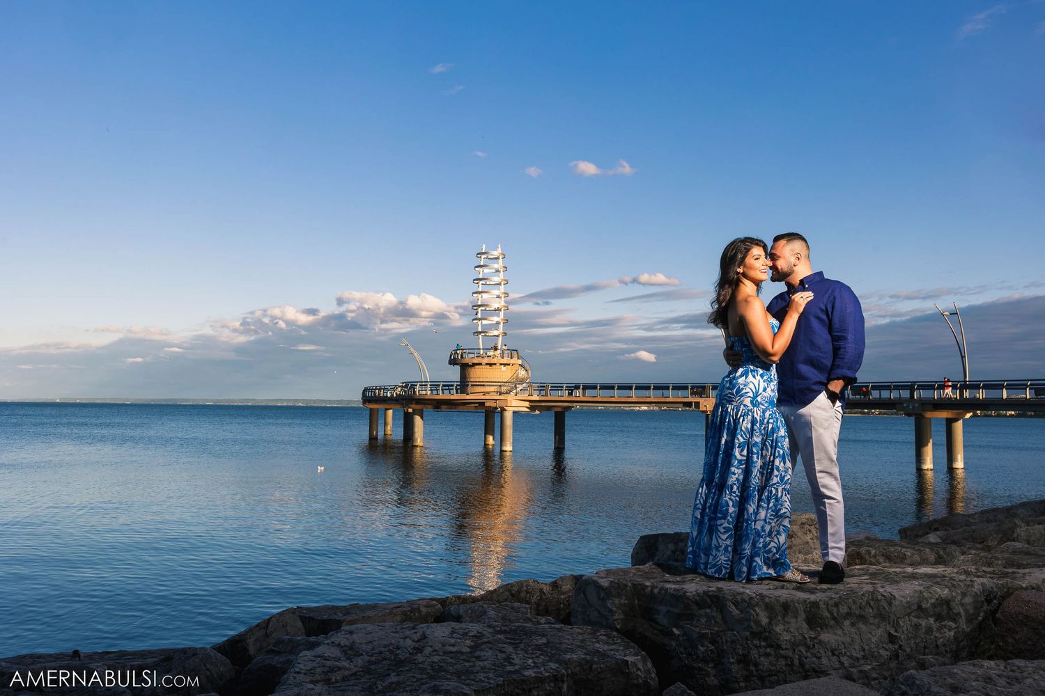 Couple embracing on rocky shore with scenic pier and ocean view at sunset.