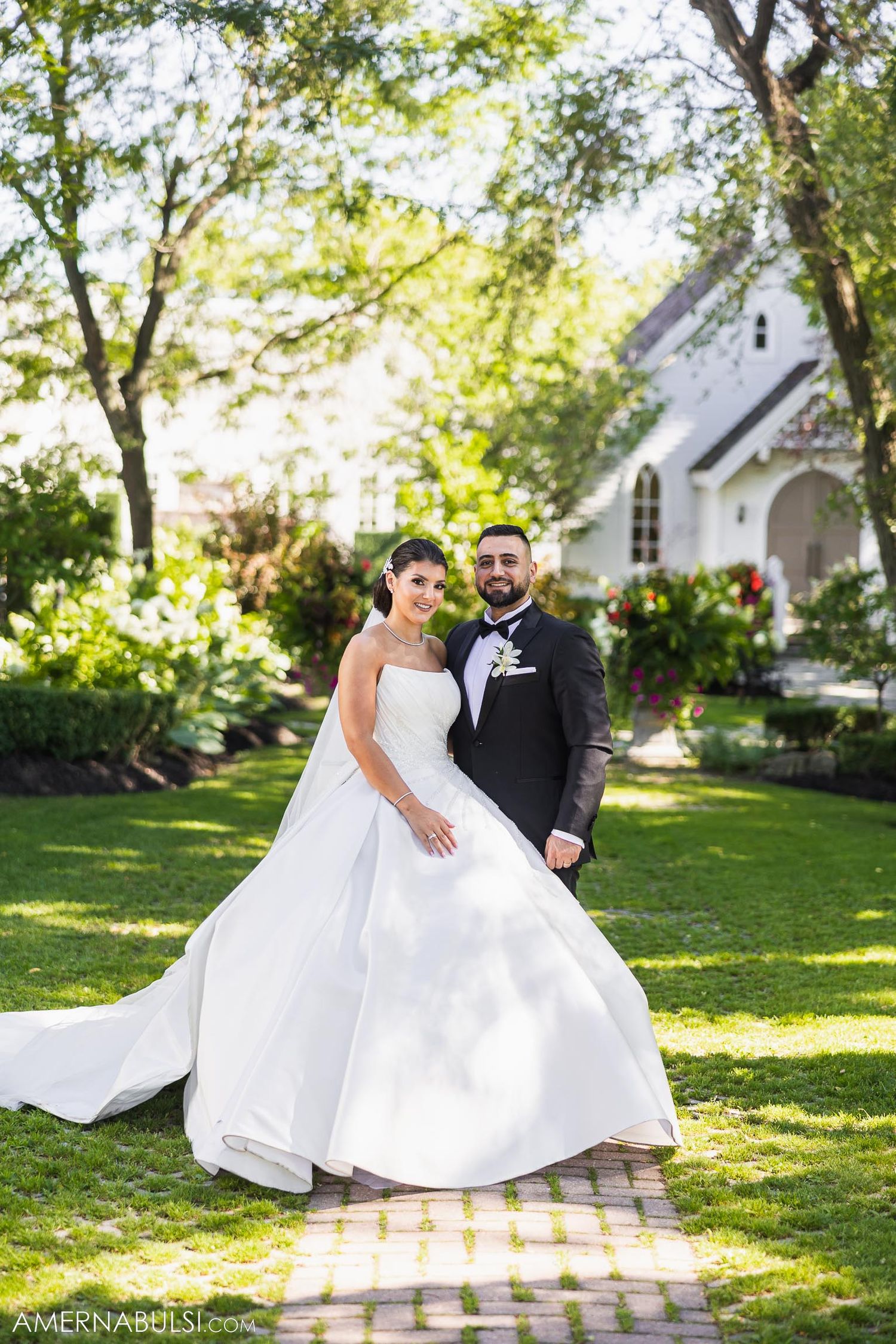 A bride and groom pose on a garden path with a white chapel in the background.