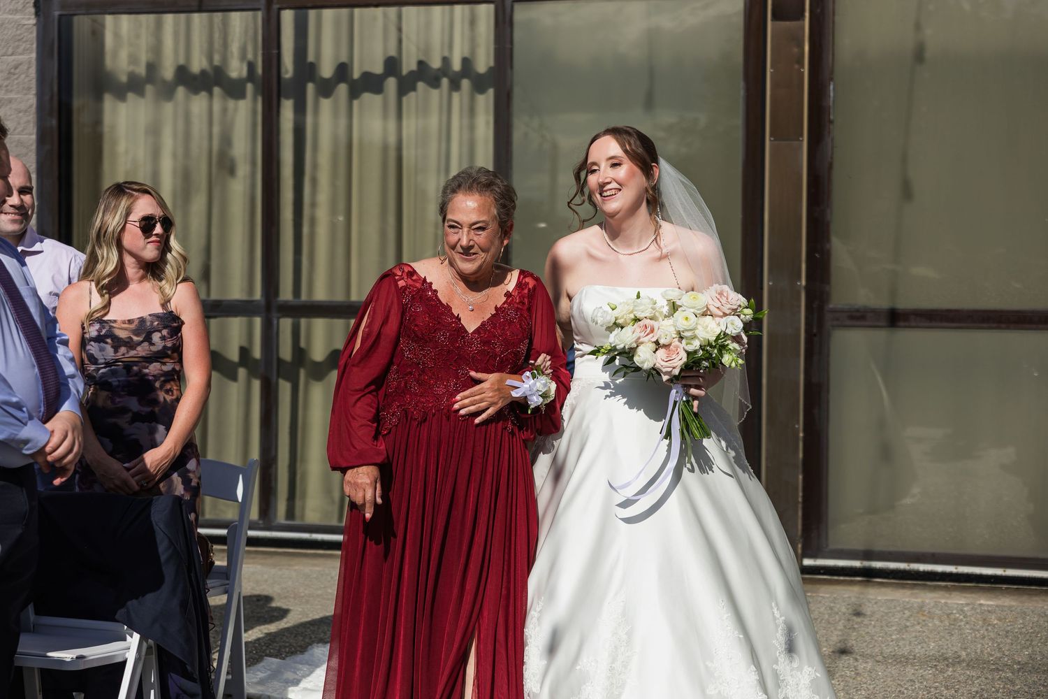 Bride in white gown walks with older woman in red dress at outdoor wedding.