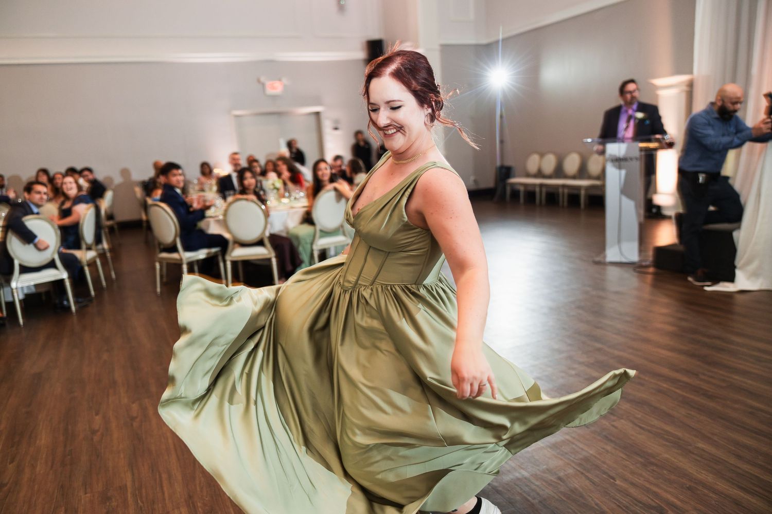Smiling bridesmaid in sage green dress twirls on dance floor at elegant wedding reception.
