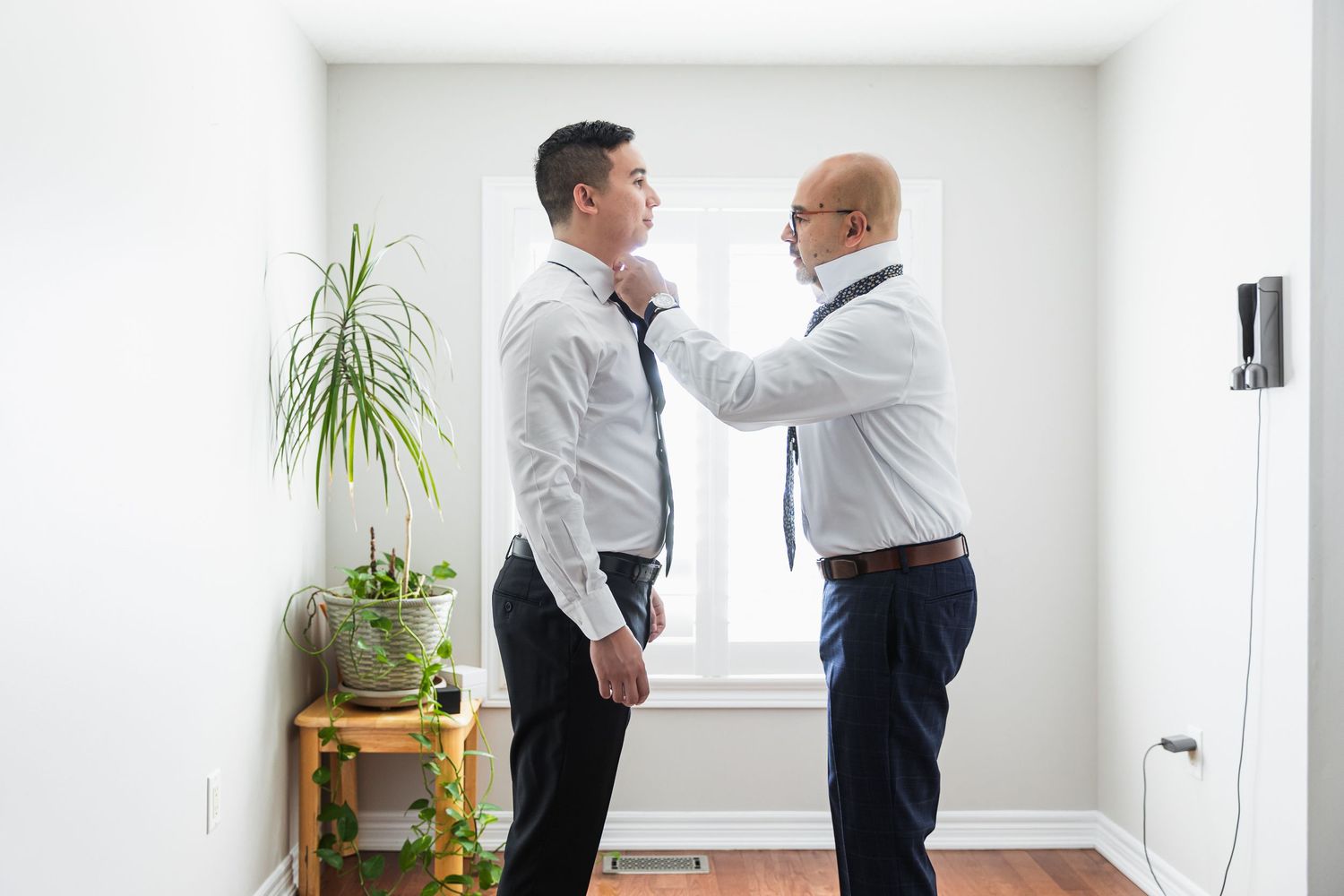 Man helping groom adjust tie while getting ready for wedding ceremony in bright white room.