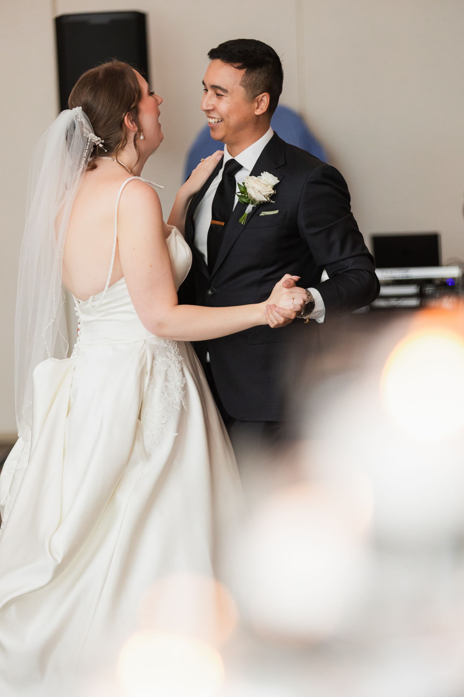 Bride and groom share their first dance, smiling at each other, bride in white gown with veil, groom in black suit.