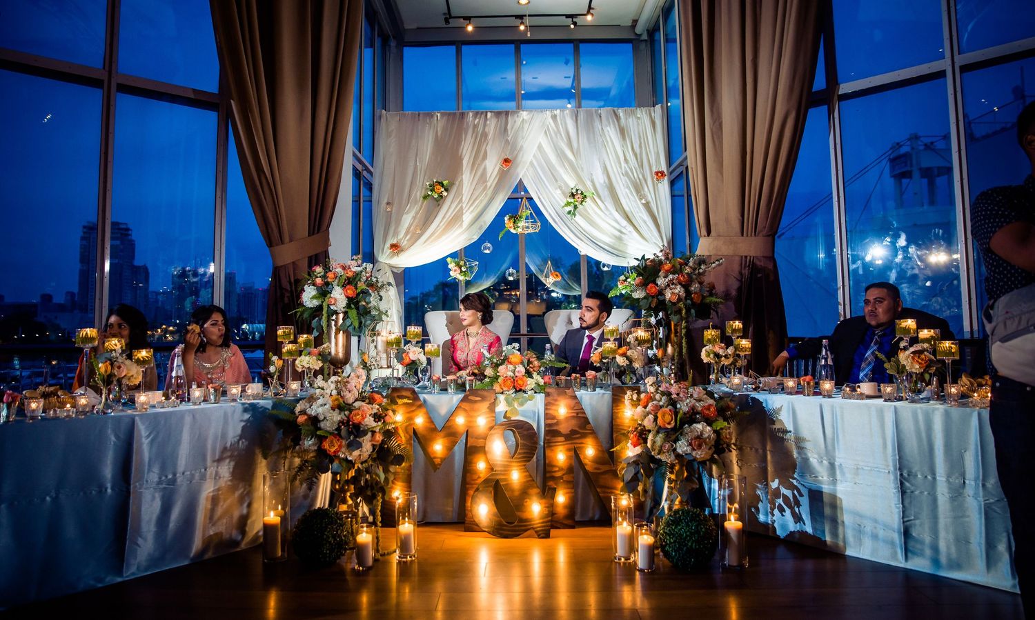 Luxurious wedding reception setup with candlelit tables and dramatic blue evening lighting through large windows.