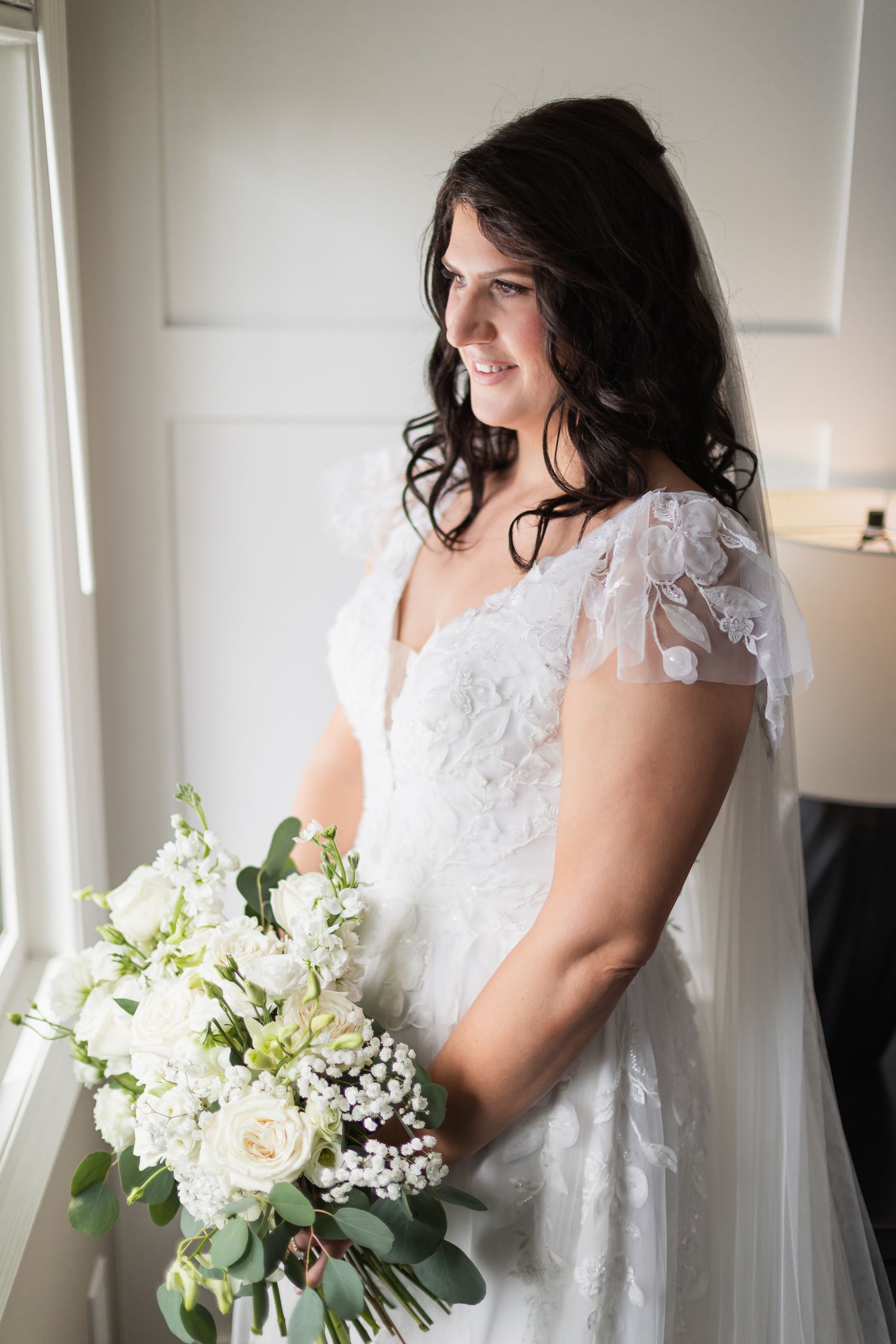 Smiling bride in lace cap-sleeve wedding gown holding white rose bouquet near bright window.