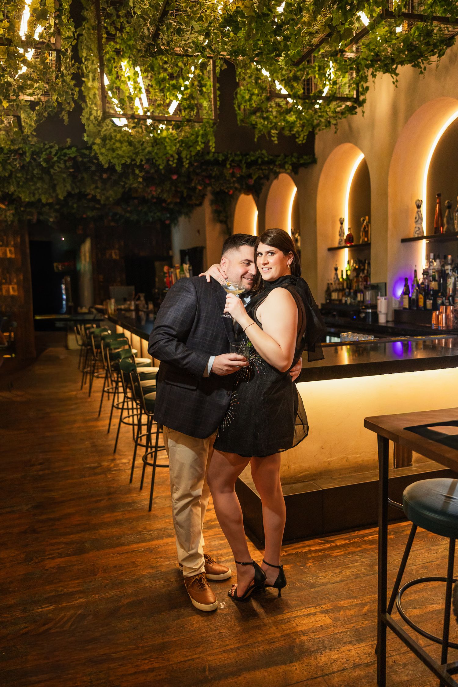 Couple embracing at an upscale bar with green vine ceiling decor, warm arched lighting, and a long wooden counter.