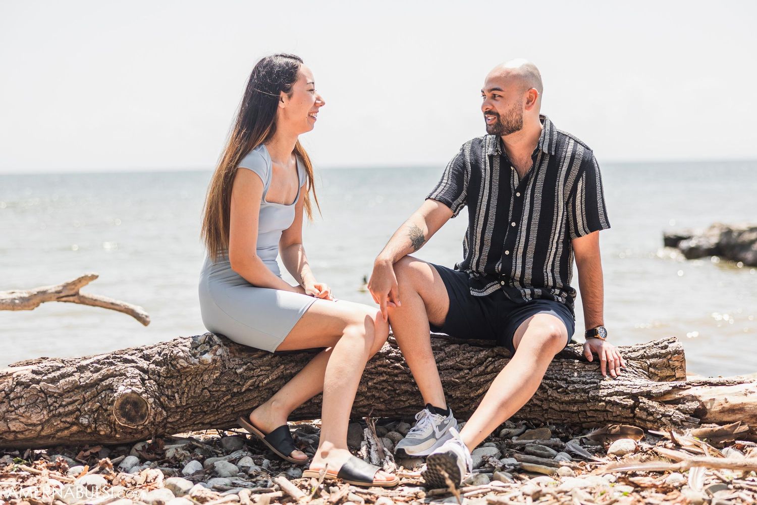A couple sits together on a fallen log at the beach on a sunny day.