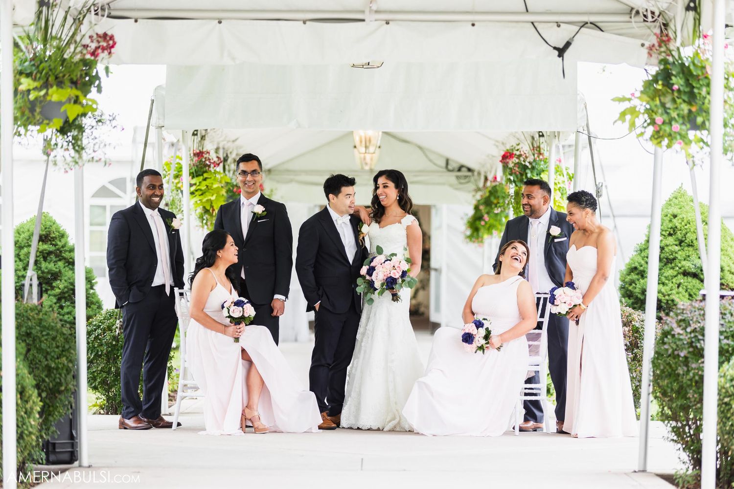 Wedding party poses under white tent with hanging plants and elegant decor.