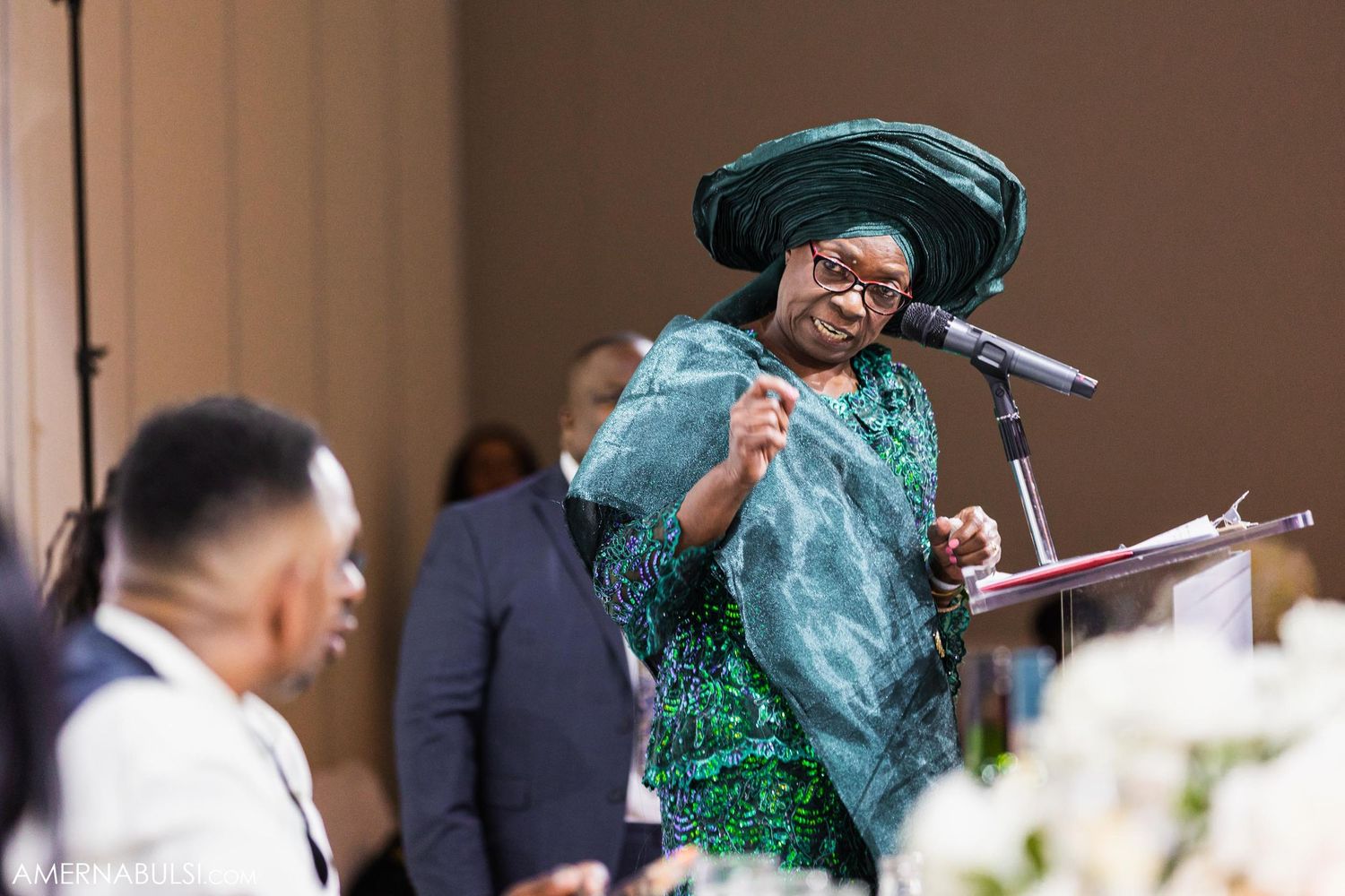 Guest in turquoise attire enthusiastically speaks at microphone during wedding reception.
