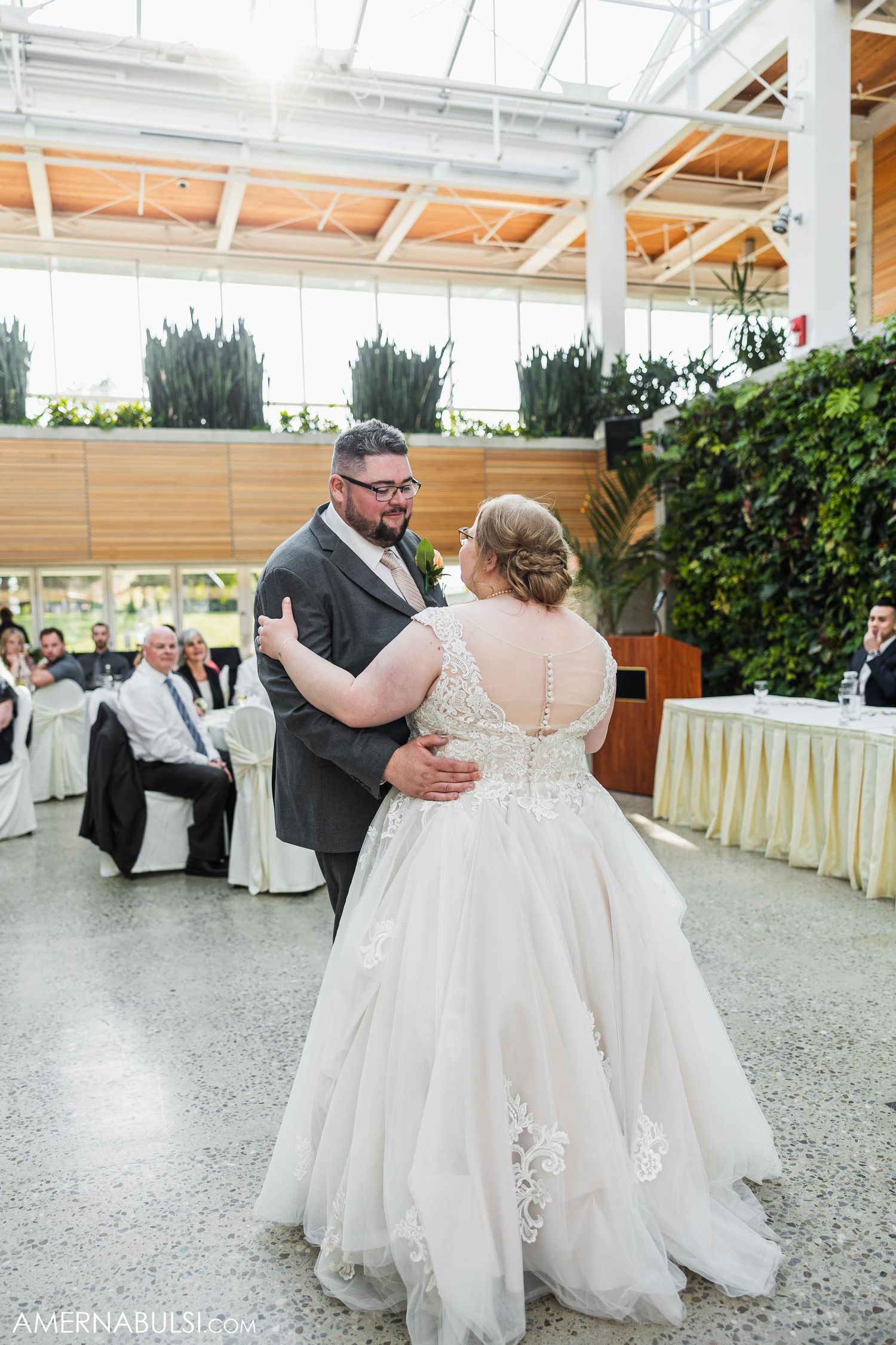 A couple embraces while dancing at their wedding reception with guests seated in the background.