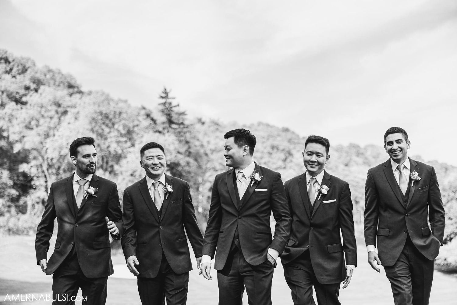 Groomsmen in dark suits walk together laughing in black and white photo.