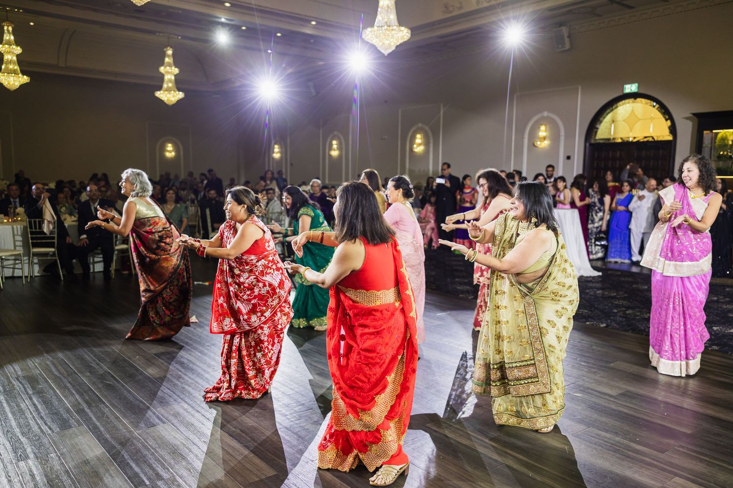 Guests in colorful traditional saris dance together on a wooden floor at an elegant reception venue.