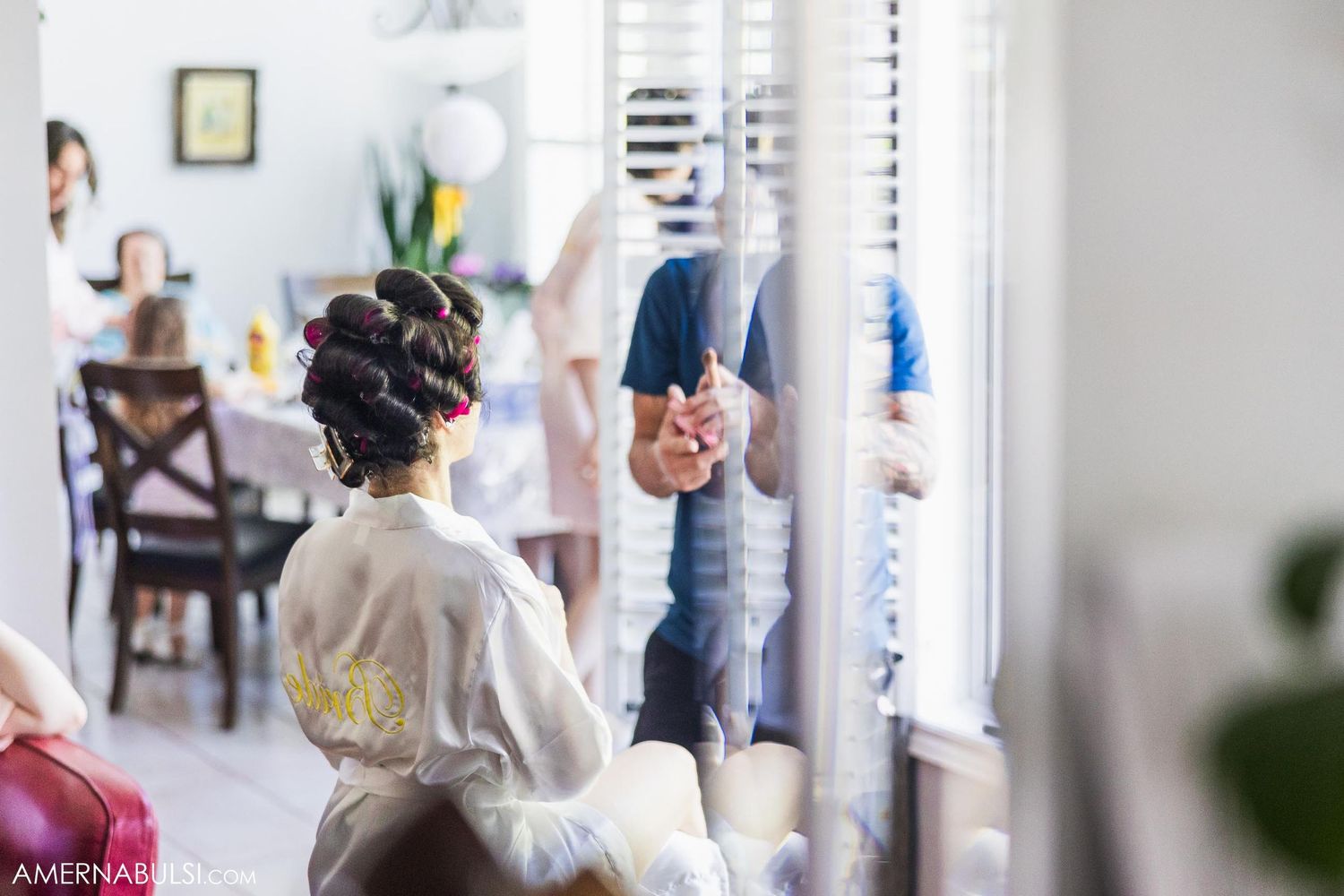 Someone in a robe sits at a window while others prepare in the background of a bright room.