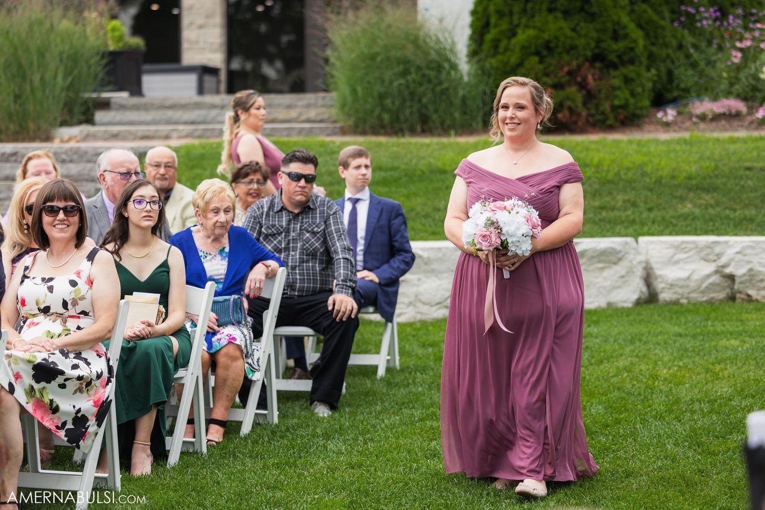 Bridesmaid in mauve dress walks down aisle past seated wedding guests during outdoor ceremony.