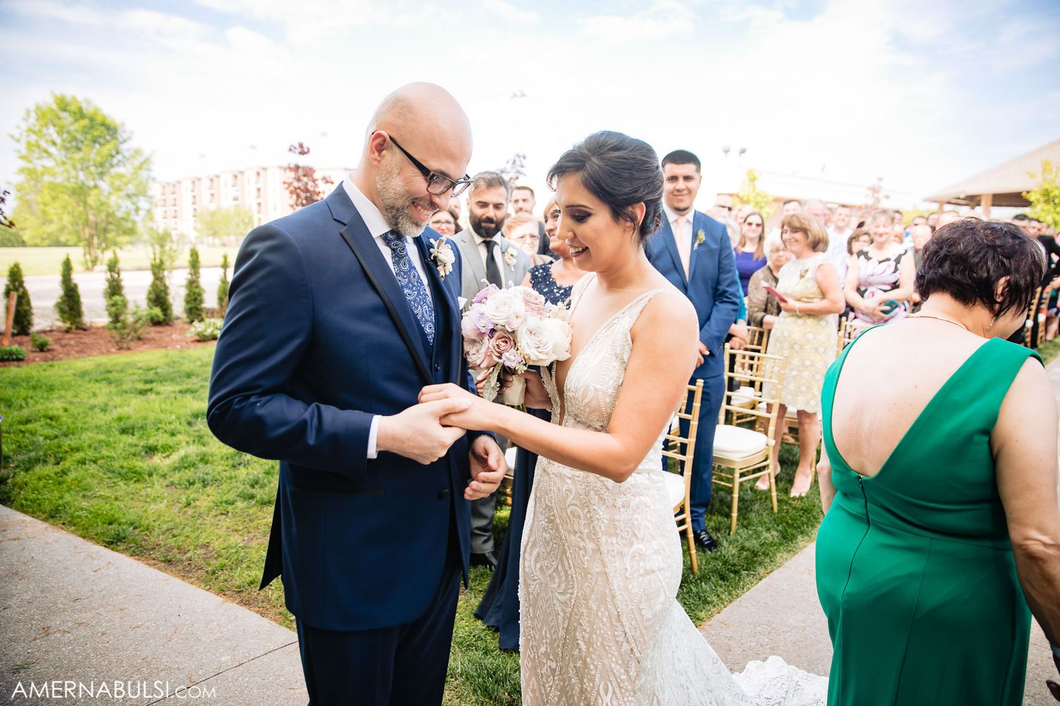 Outdoor wedding ceremony with guests gathered on a sunny day with green landscaping.