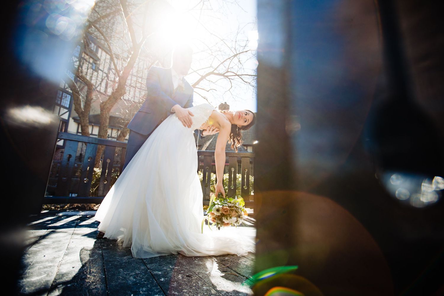 Newlyweds share a romantic moment in sunlit courtyard with lens flare and dramatic lighting.