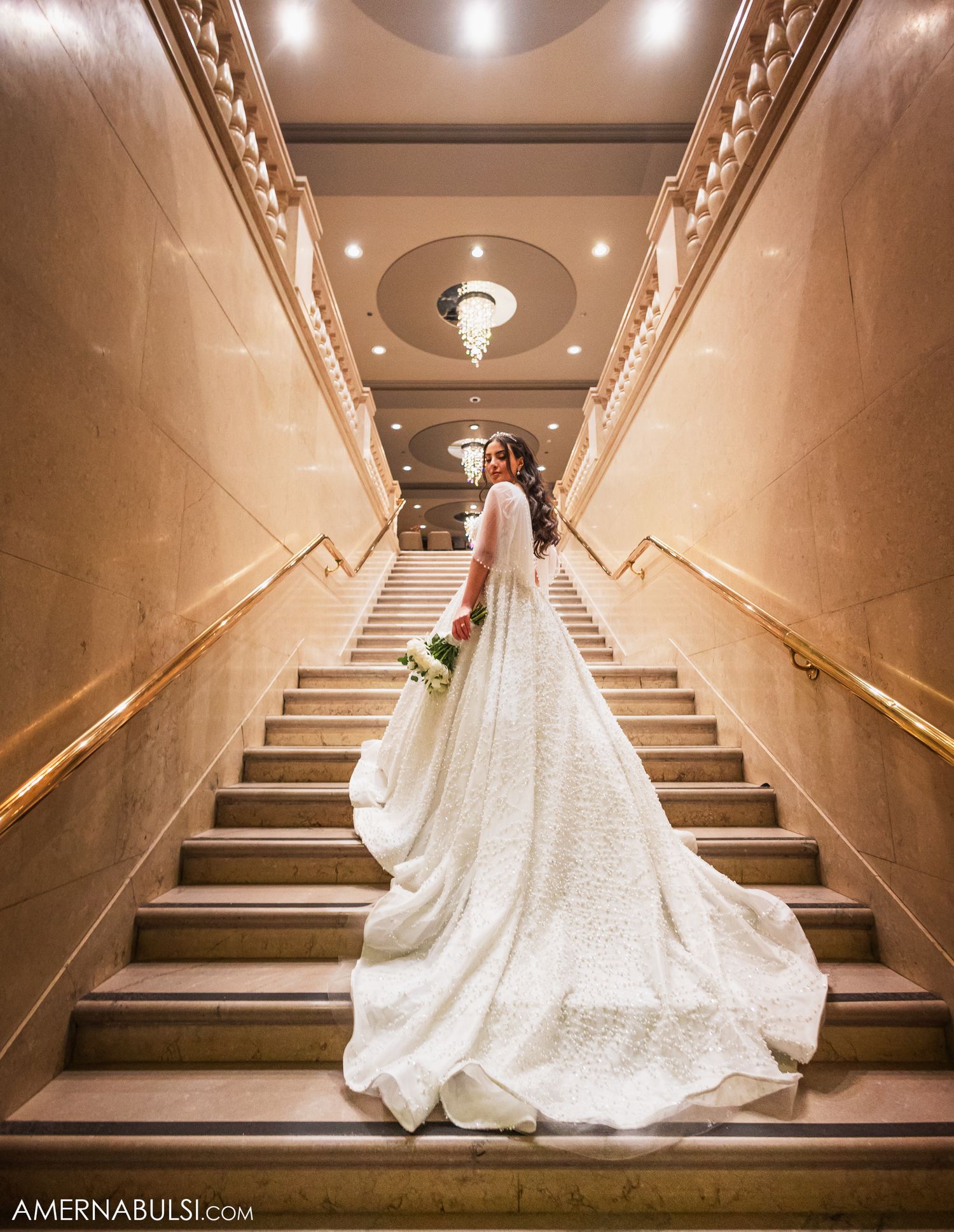 A flowing white wedding gown with a dramatic train cascades down an elegant marble staircase in warm lighting.