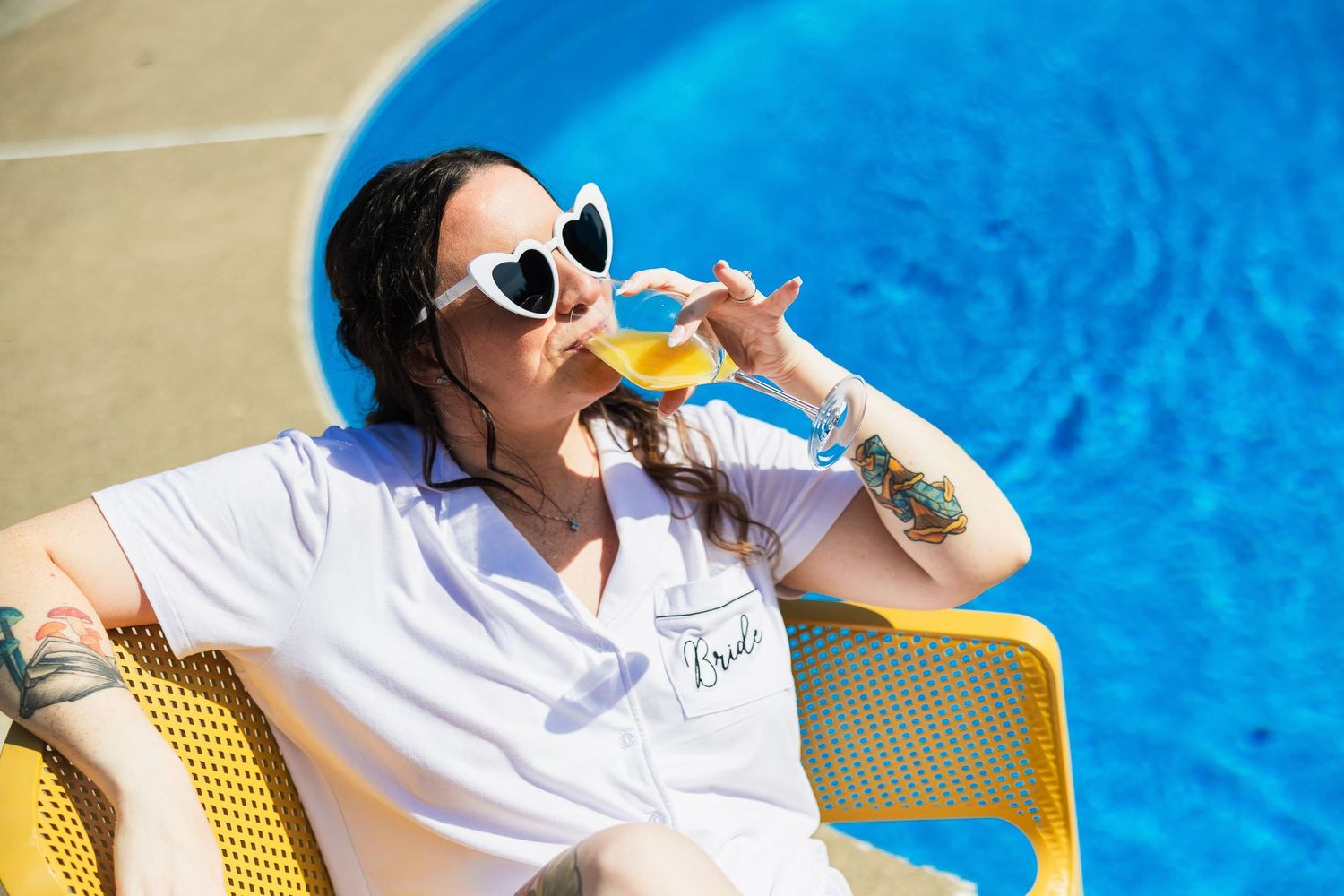 Person in white robe relaxing on yellow chair by pool while sipping a drink.