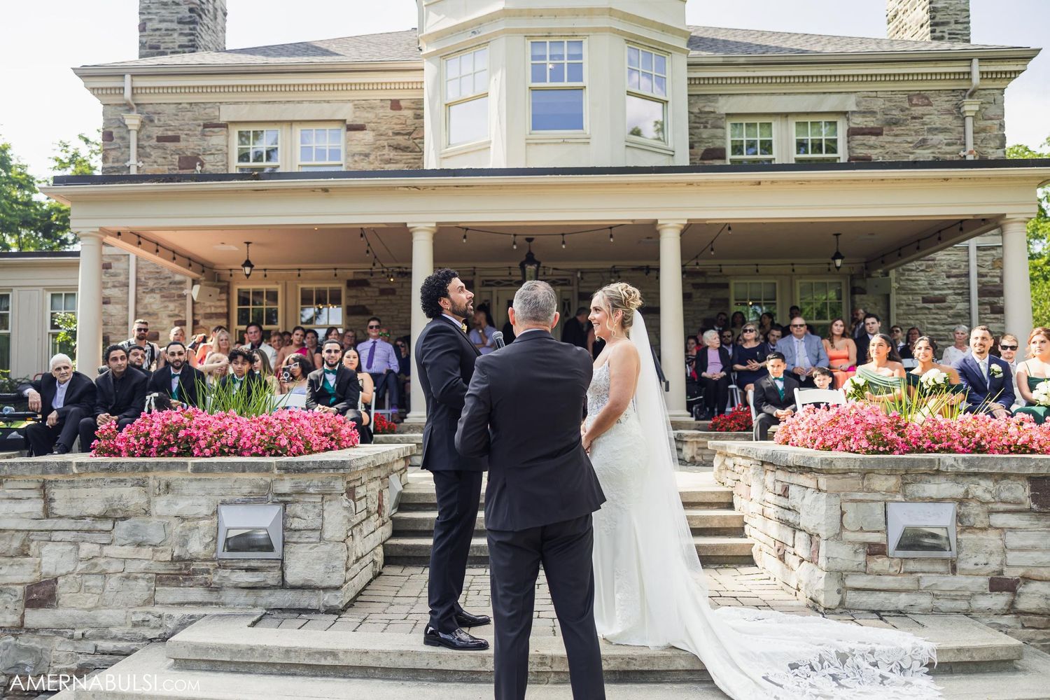 Outdoor wedding ceremony in front of a grand stone mansion with pink flowers decorating the entrance.