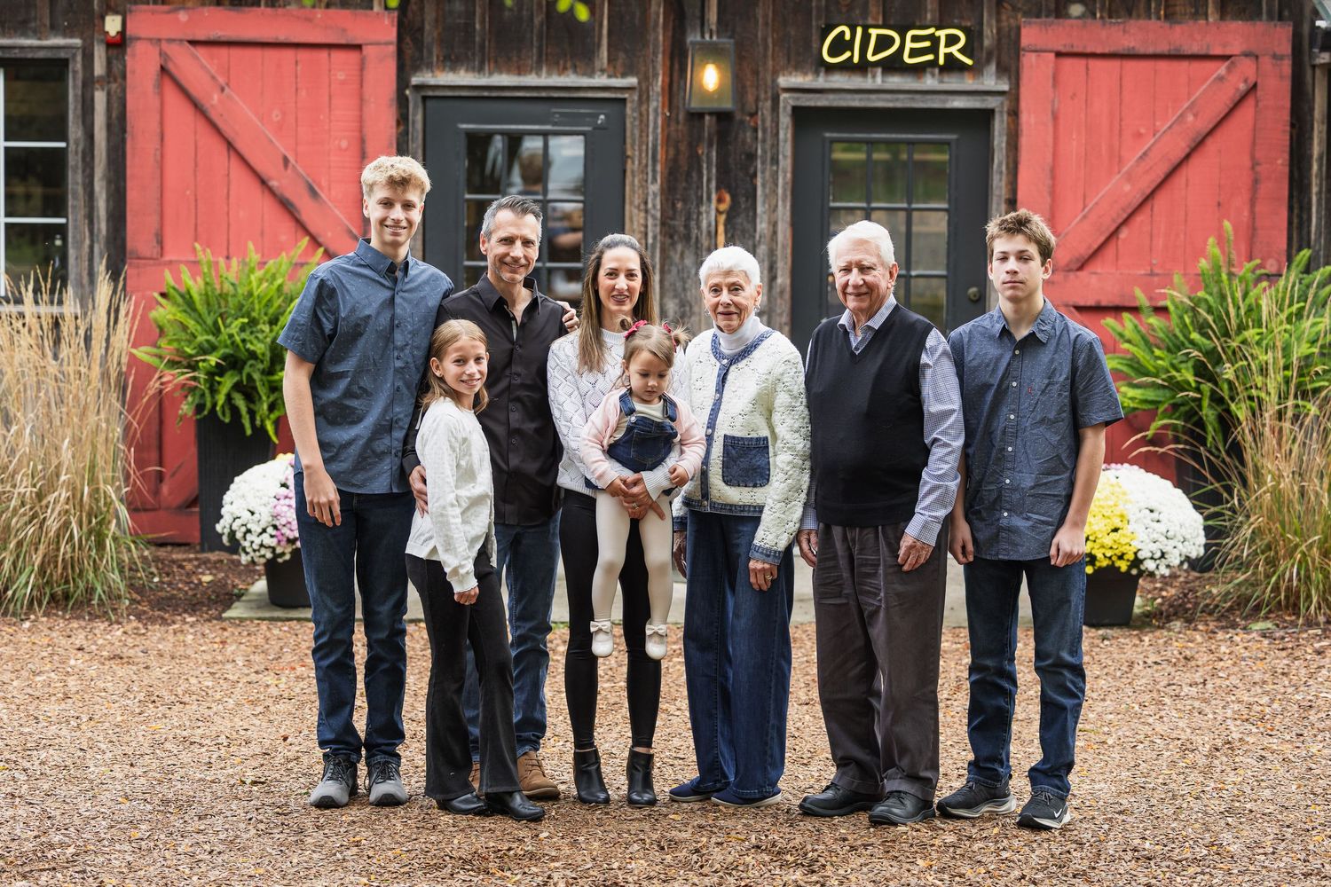 A group of people stand together in front of a rustic red barn with a cider sign on a fall day.
