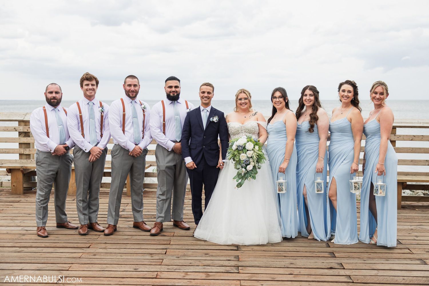 Wedding party in light blue dresses and gray suits poses on wooden boardwalk by the ocean.