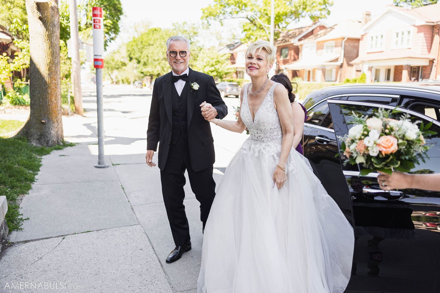 Couple walking arm in arm on sidewalk next to decorated wedding car.
