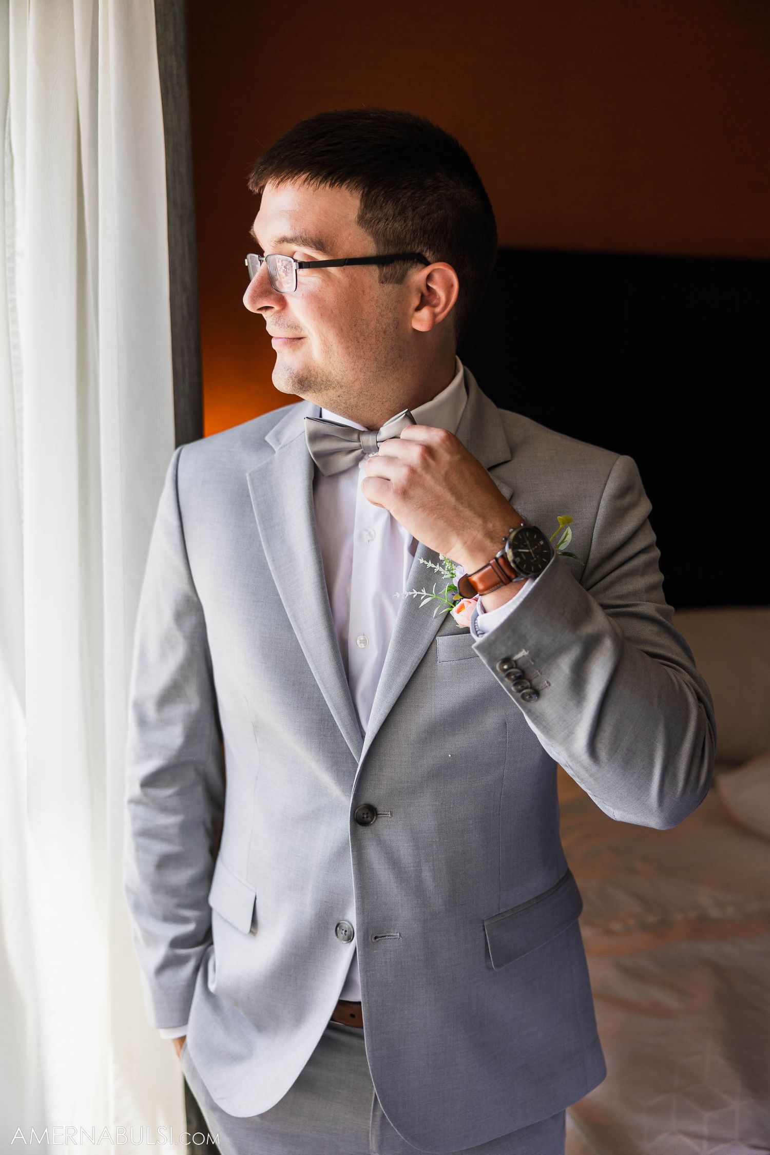 Person in light gray suit adjusts bowtie while standing by window.