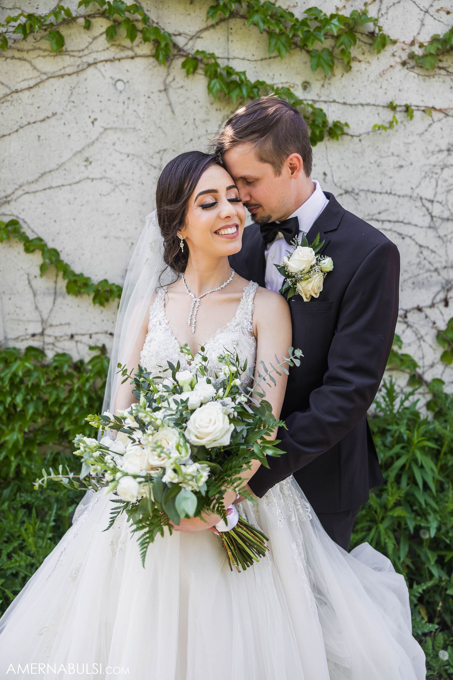 Elegant bride holds white floral bouquet while sharing intimate moment against ivy-covered wall.