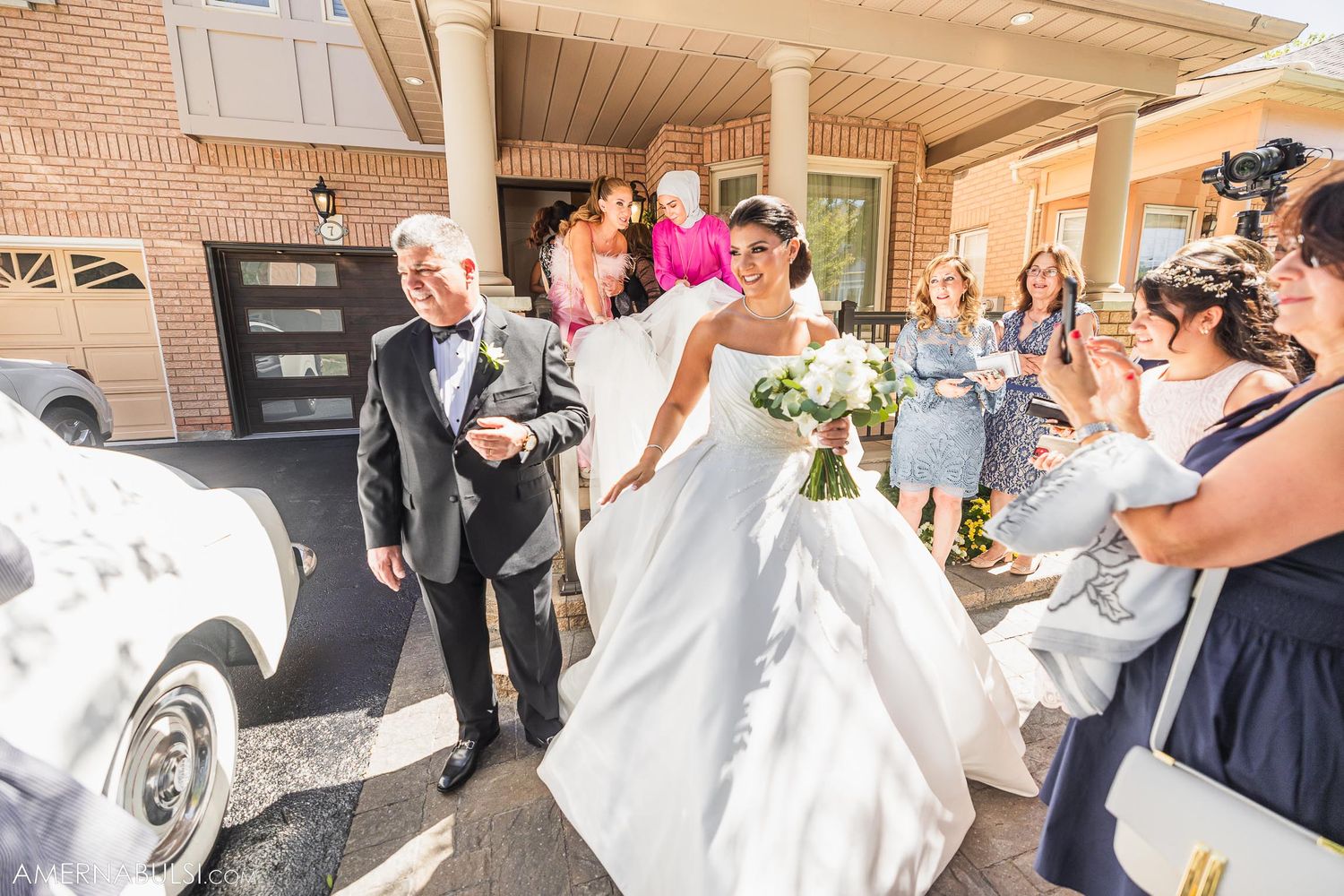 Wedding party helping bride exit vintage car outside brick building.