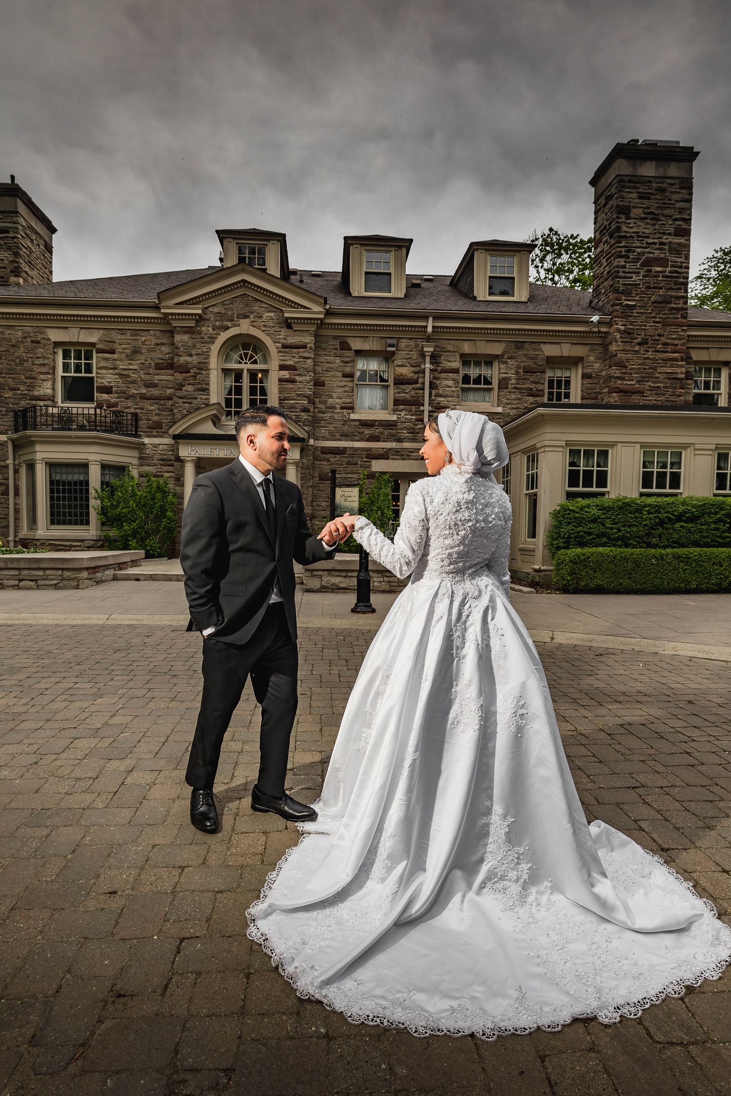 Bride and groom pose playfully in front of elegant stone mansion with dramatic cloudy sky.