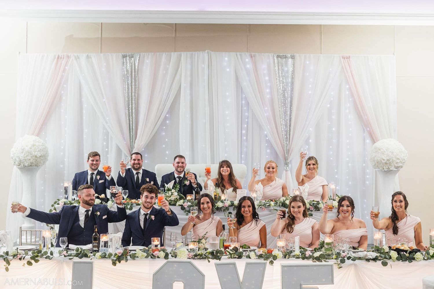 Wedding party poses together at head table with elegant white drapery backdrop.