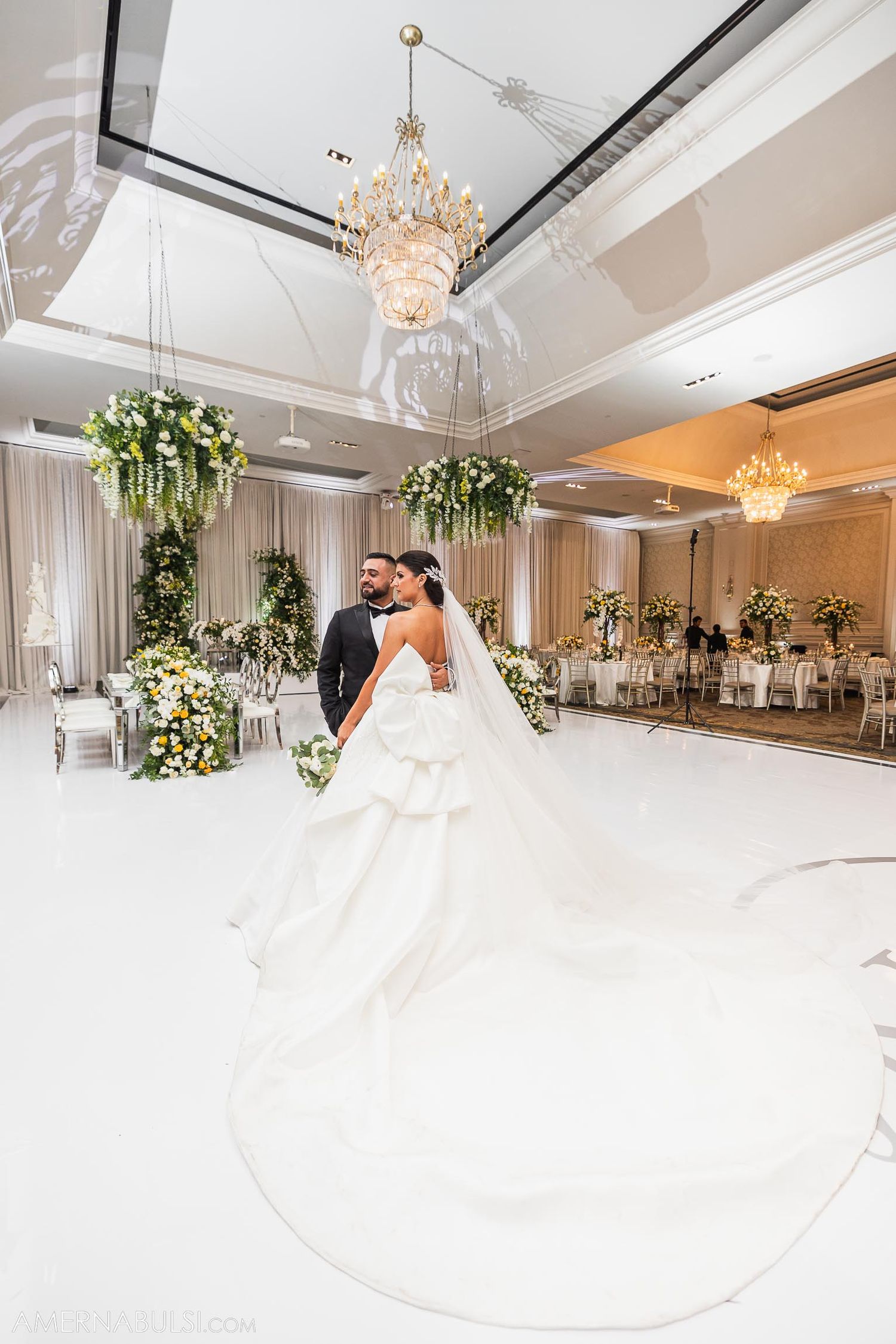 Elegant wedding couple dancing in luxurious ballroom decorated with floral arrangements and crystal chandeliers.