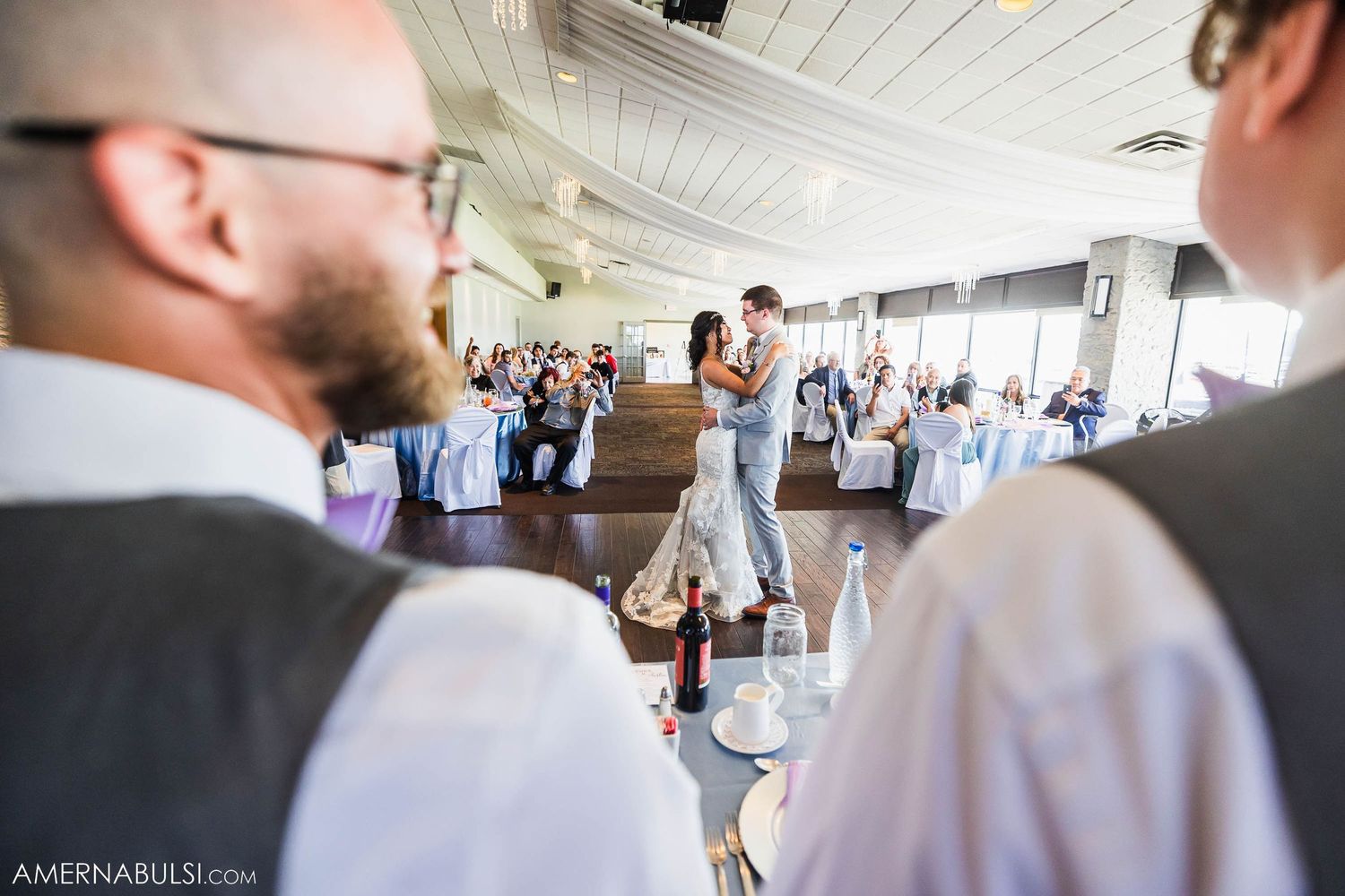 Guests observe a couple dancing in an elegant reception venue with white draped ceilings.