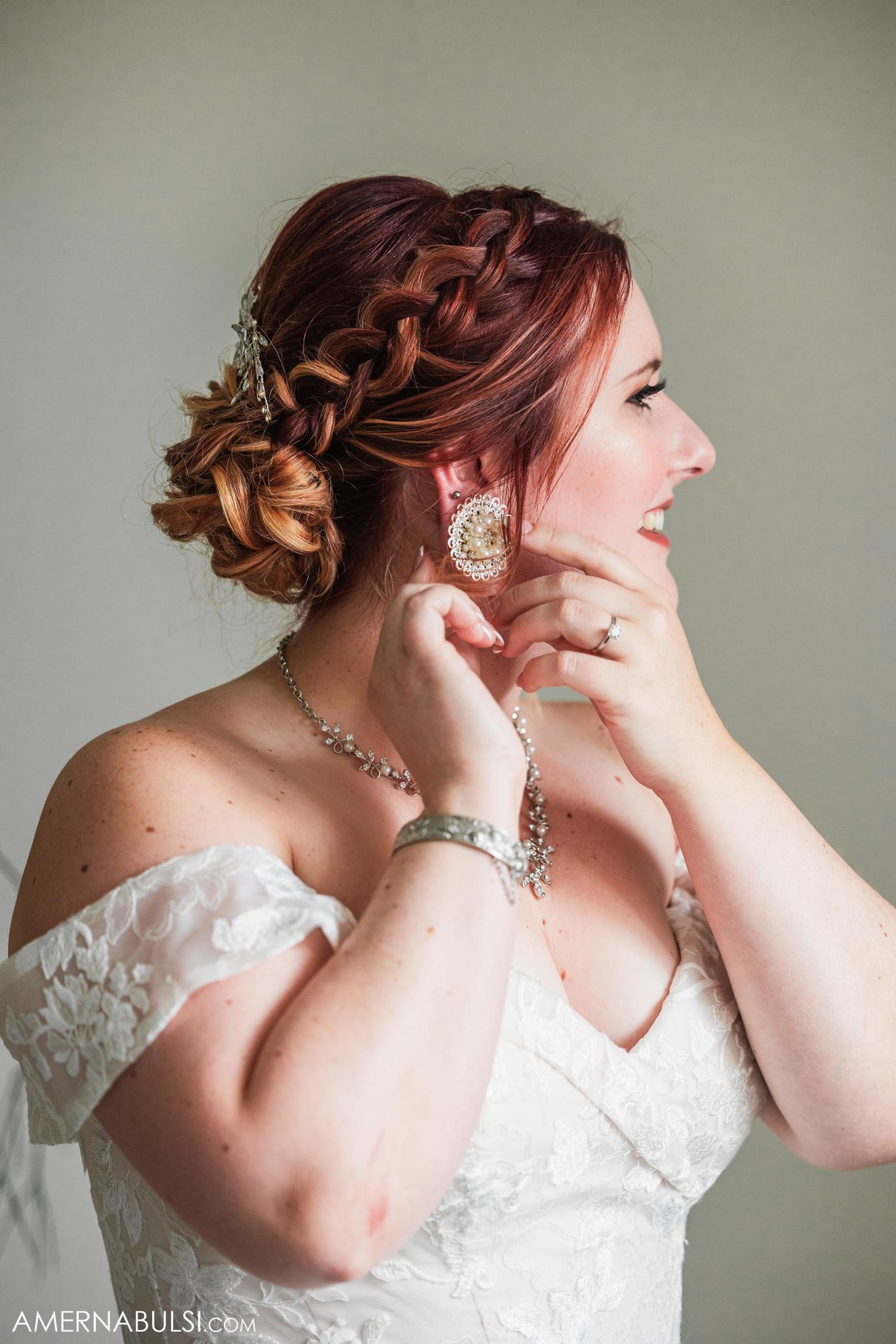 Bride with ornate braided updo wearing white gown and jewelry smiles while adjusting earrings.