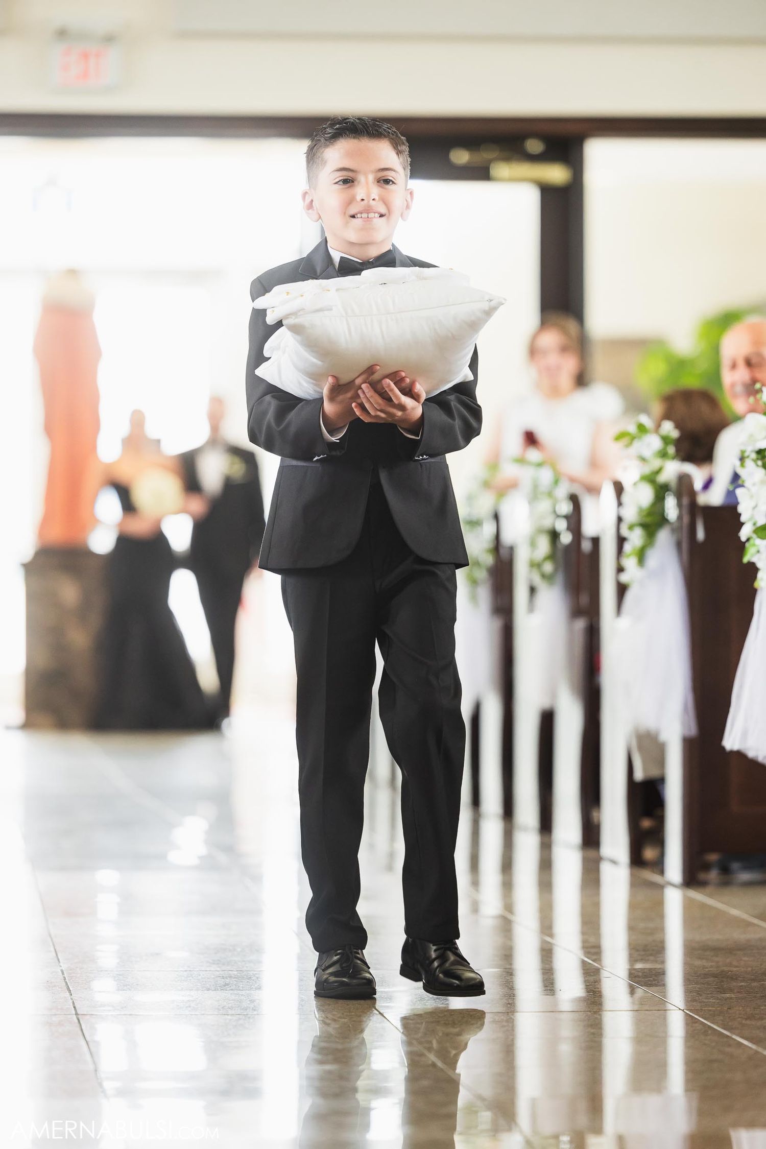Ring bearer in black suit walking down aisle carrying white pillow.