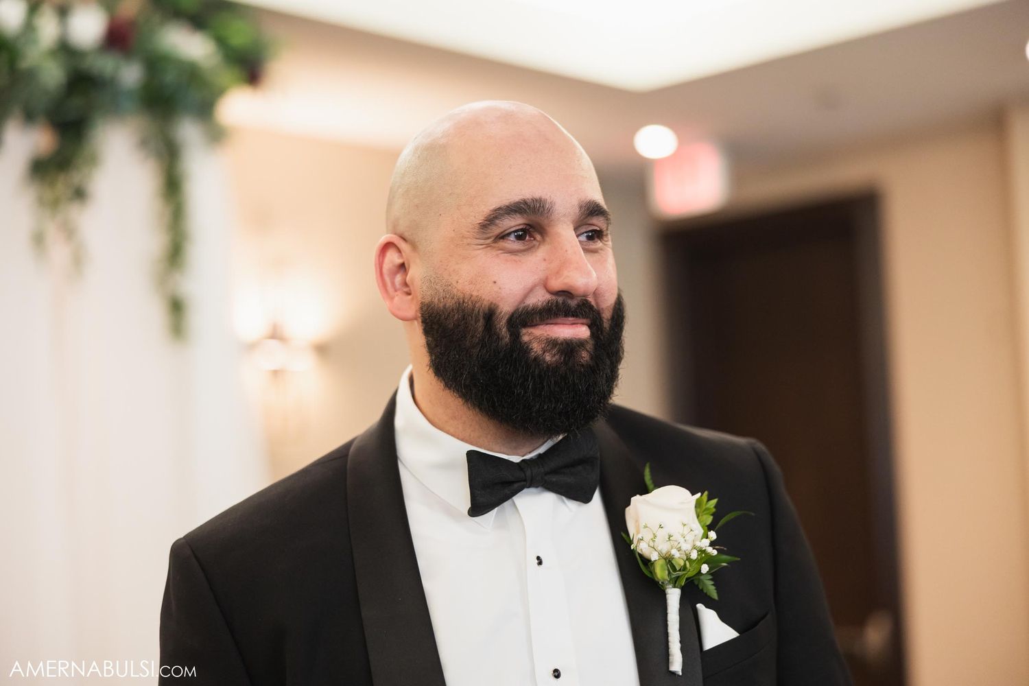 Elegant groom in black tuxedo with white boutonniere smiles during wedding celebration.