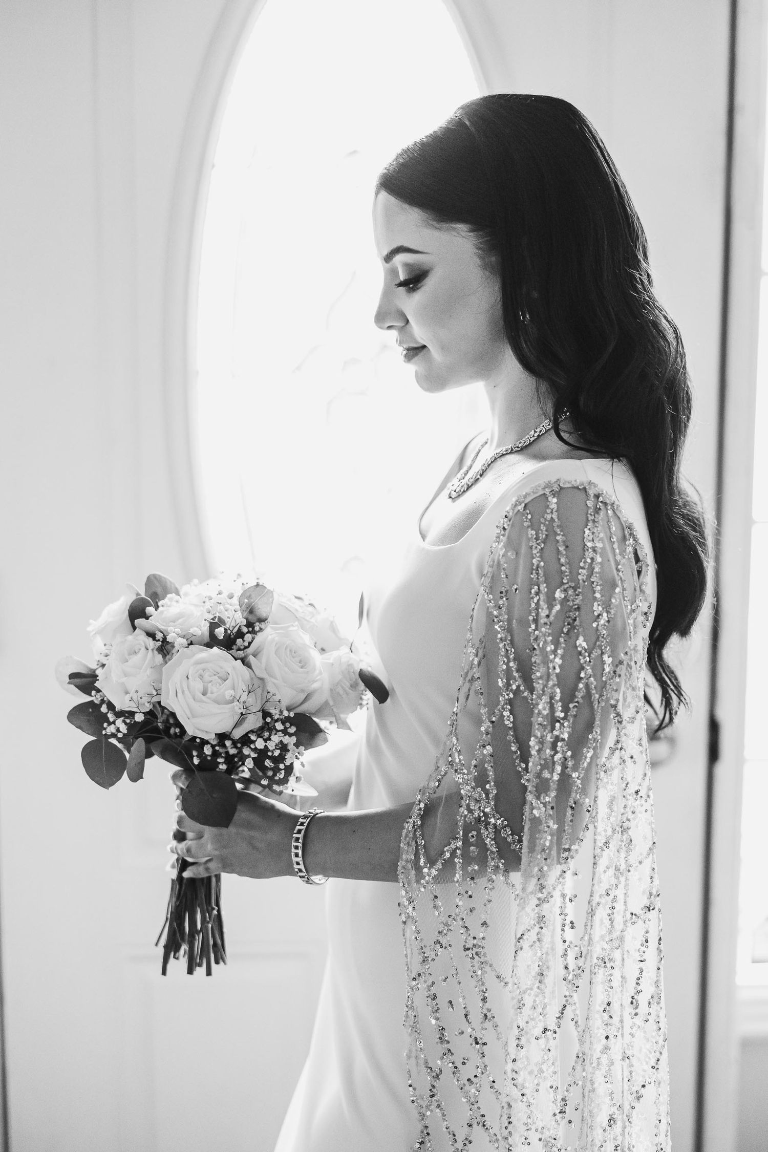 Black and white portrait of bride in sparkly gown holding white rose bouquet near window.