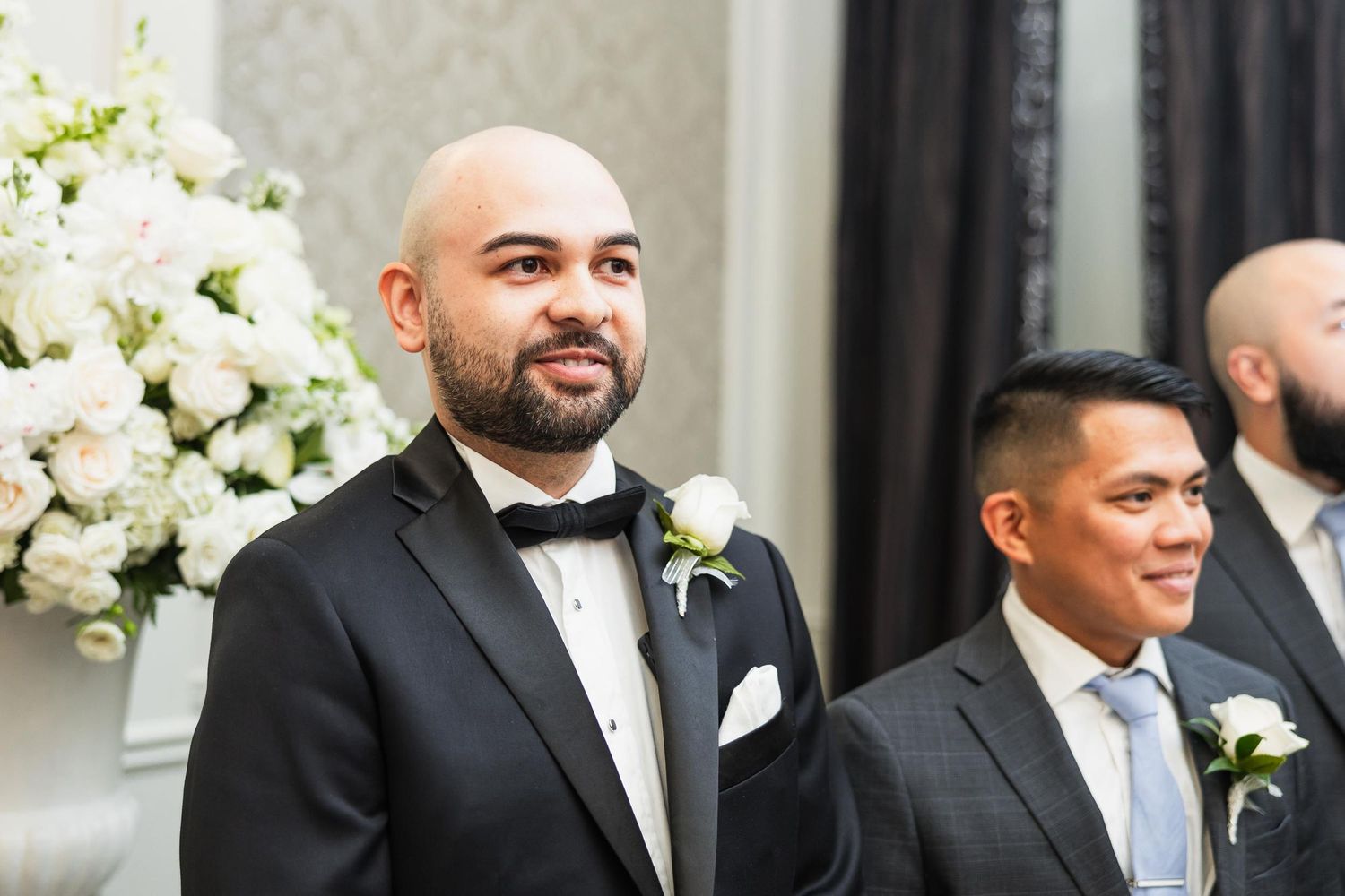 Groomsmen stand at altar with white floral arrangements during formal wedding ceremony.