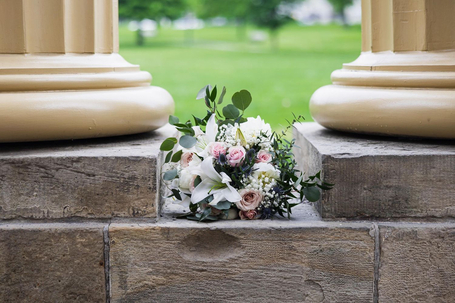 Wedding bouquet with pink roses and greenery resting on stone steps between classic columns.