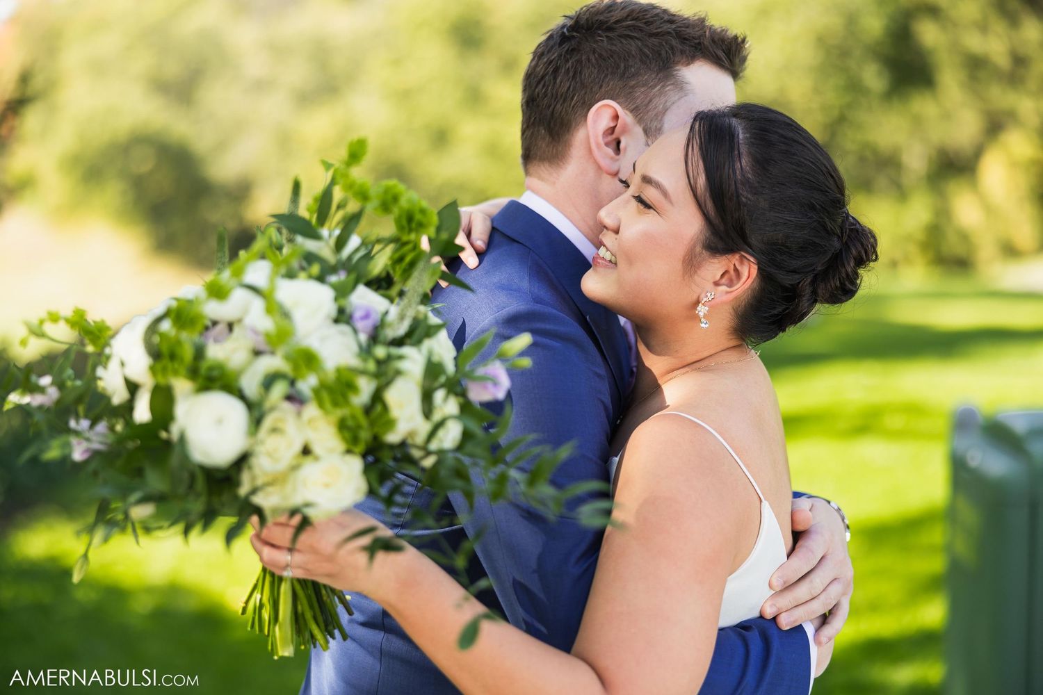 Newlyweds embrace in a sun-drenched garden setting while holding a stunning white floral bouquet.