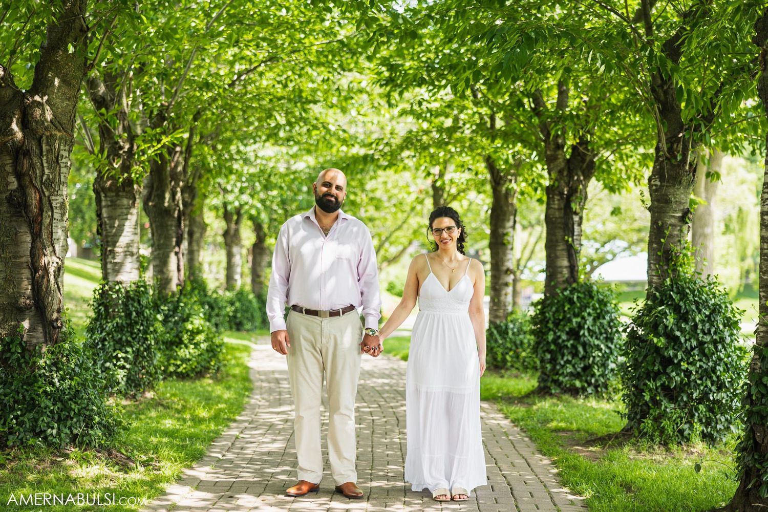 Couple walking hand in hand down tree-lined path in flowing white attire.