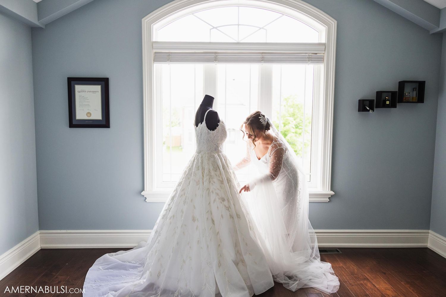 An elegant white wedding dress hangs by a bright arched window in a room with blue-gray walls.
