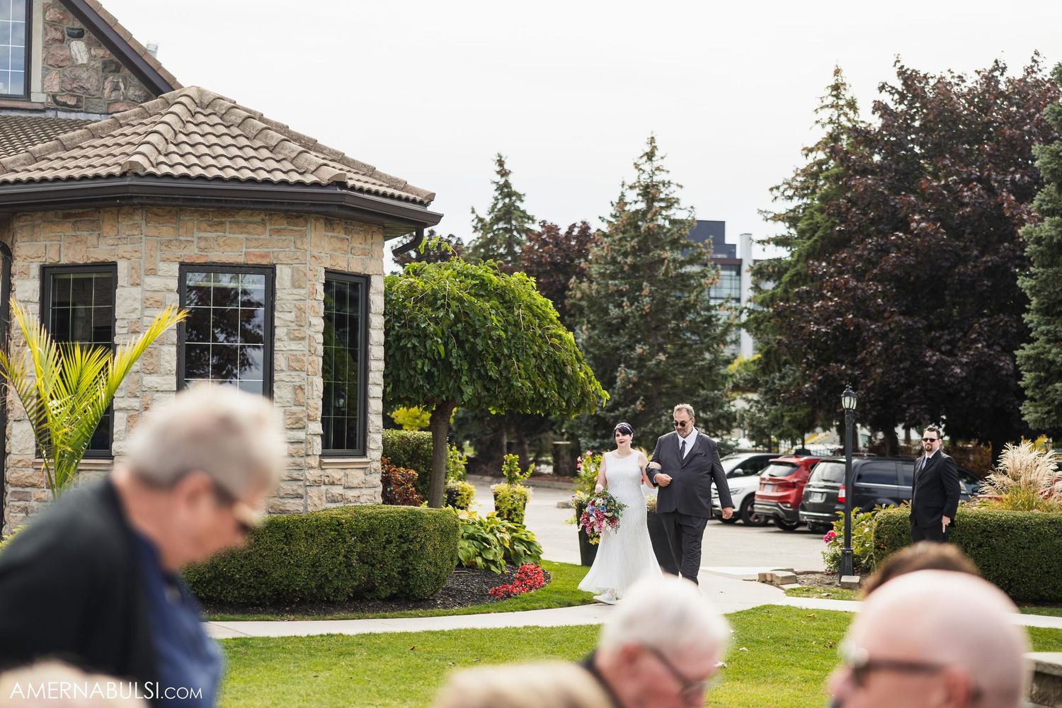 Outdoor wedding ceremony near a stone building with guests gathered on a landscaped lawn.