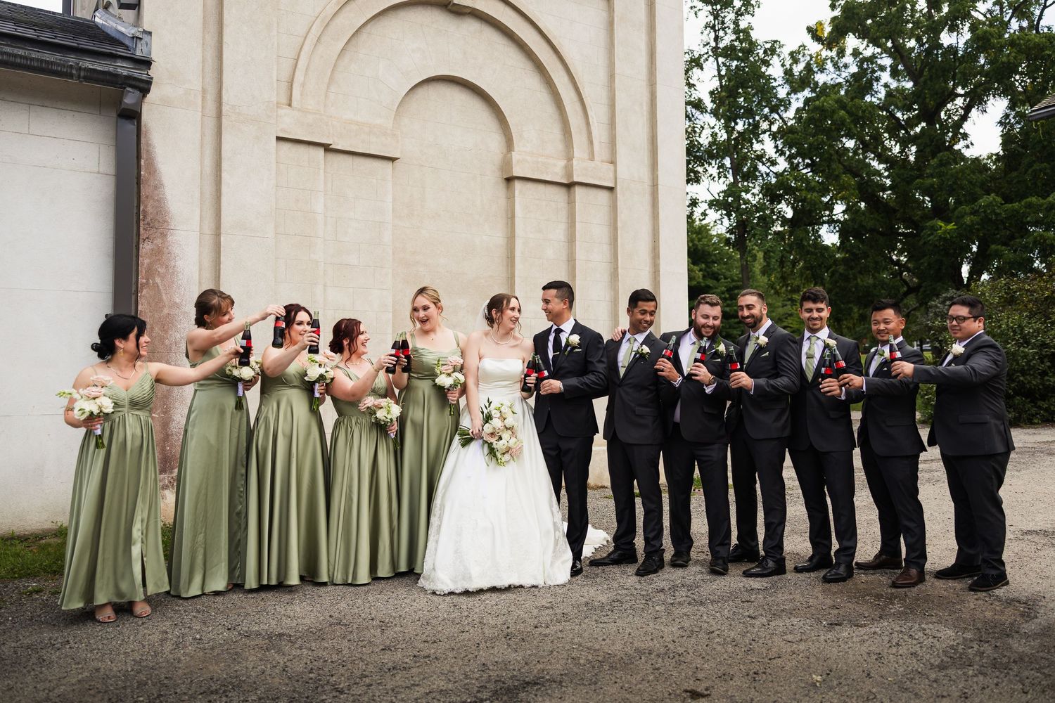 Wedding party in sage green and black poses outside stone arch building.
