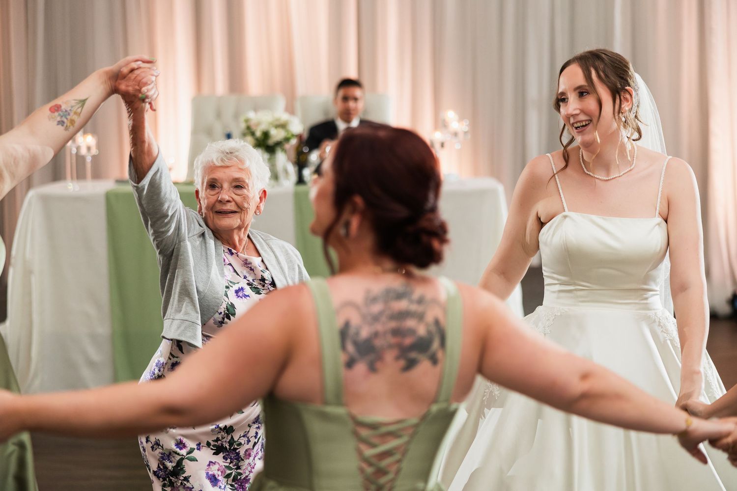 Bride in white gown dancing joyfully with elderly woman and tattooed bridesmaid at elegant wedding reception.