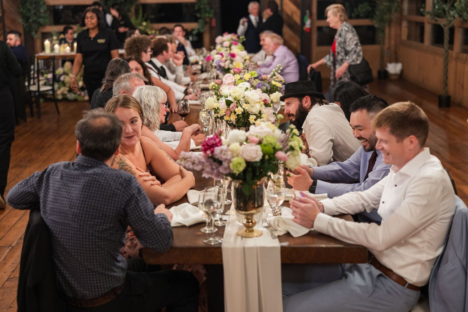 Guests seated at a long wooden table during a wedding reception, adorned with floral centerpieces in a rustic venue.