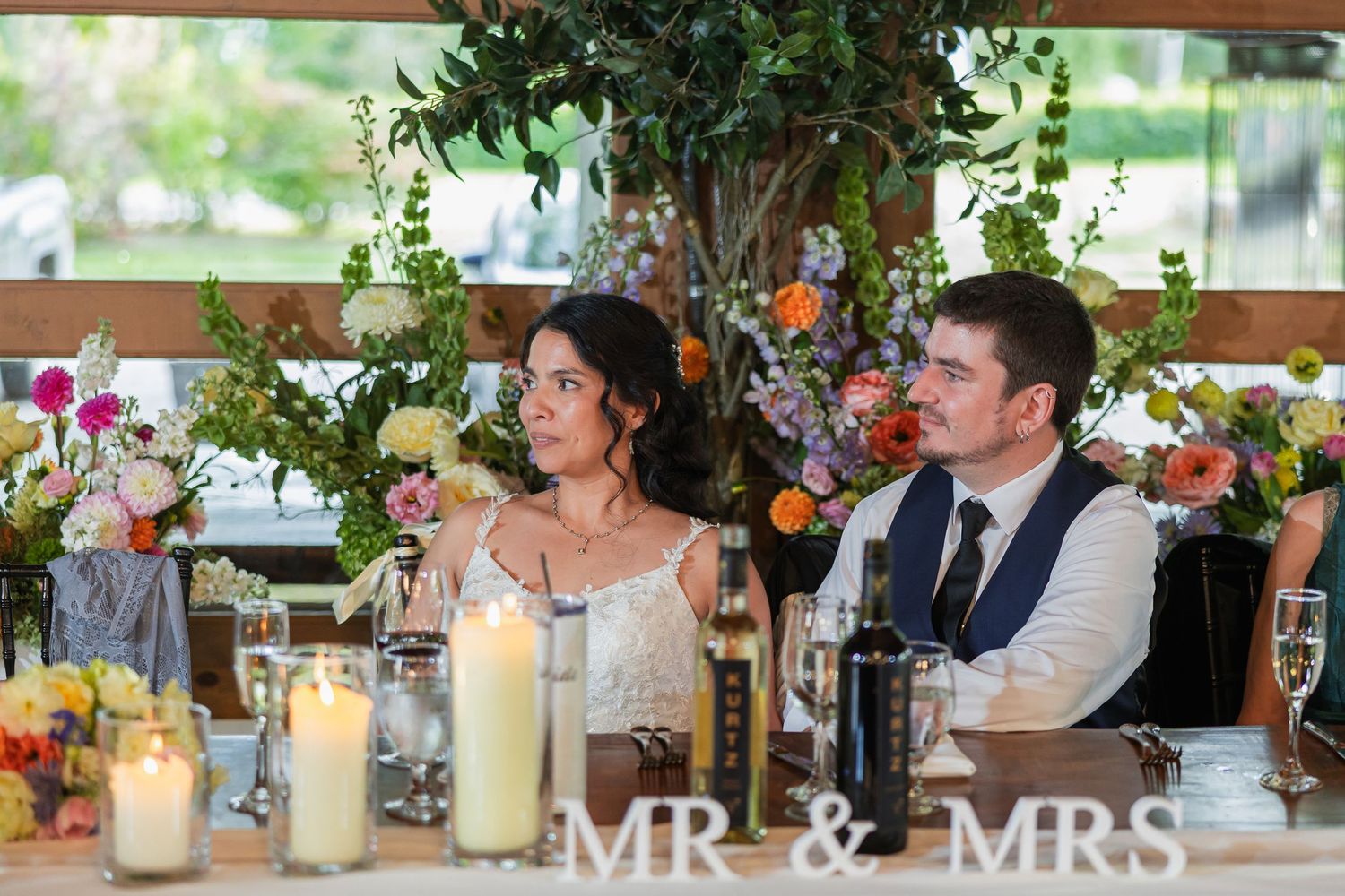 Bride and groom seated at wedding reception head table with Mr & Mrs sign, candles, and colorful floral arrangements.