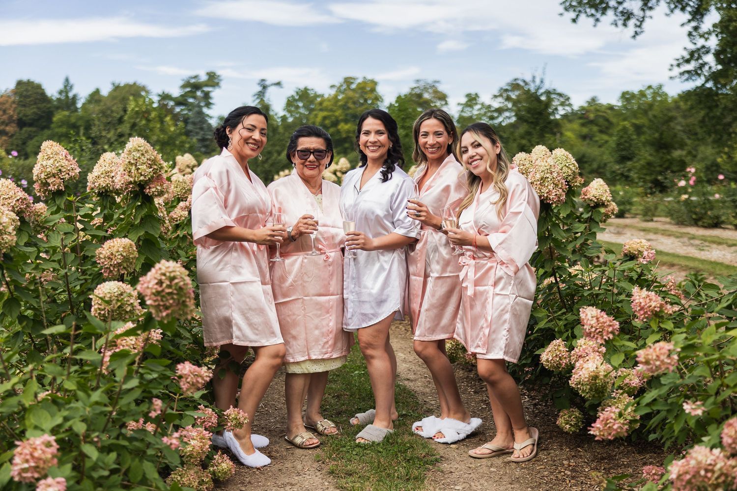 Bride and bridesmaids in matching pink robes posing among blooming hydrangeas in an outdoor garden setting.