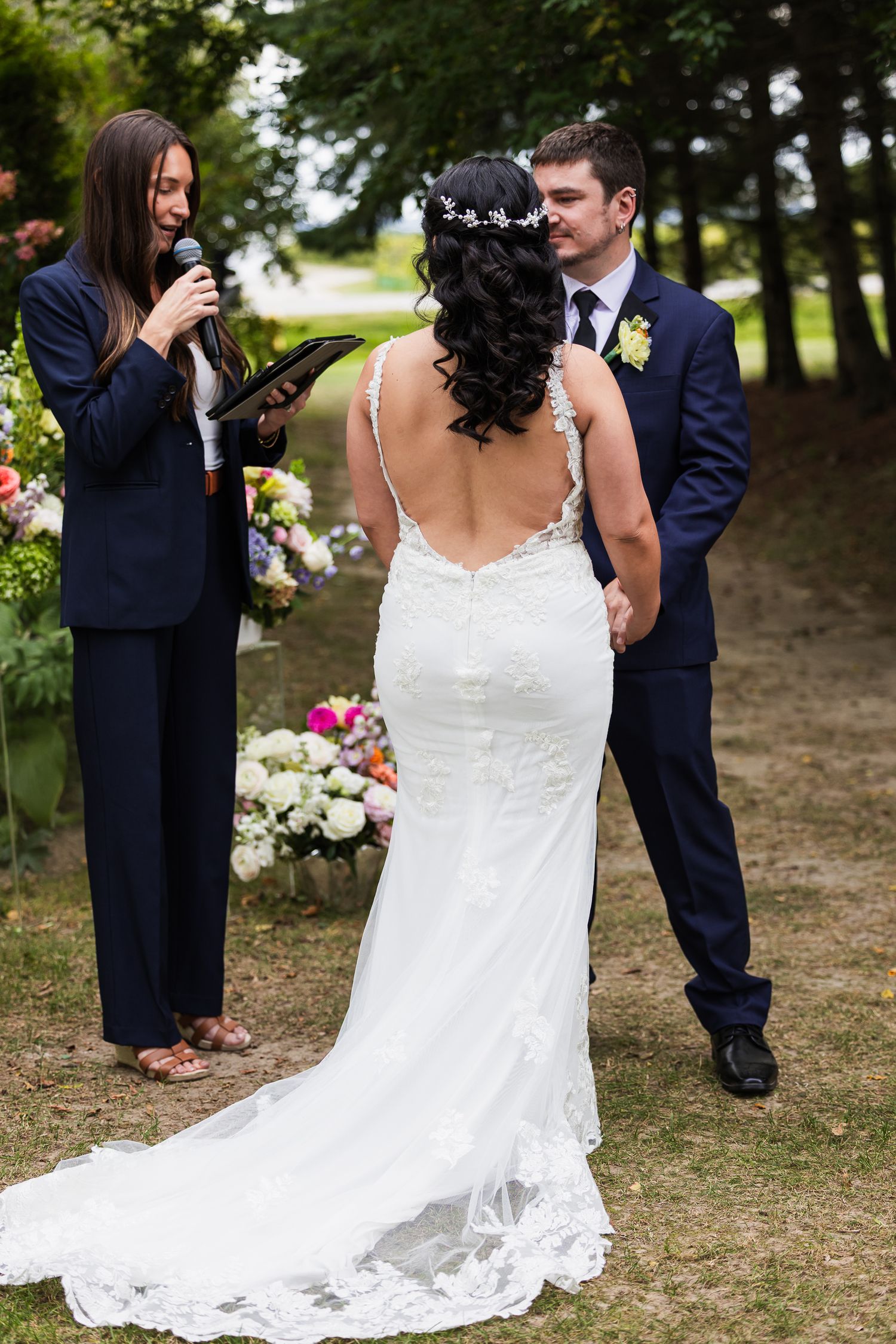 Bride and groom holding hands during outdoor wedding ceremony while officiant reads vows, surrounded by colorful floral arrangements.